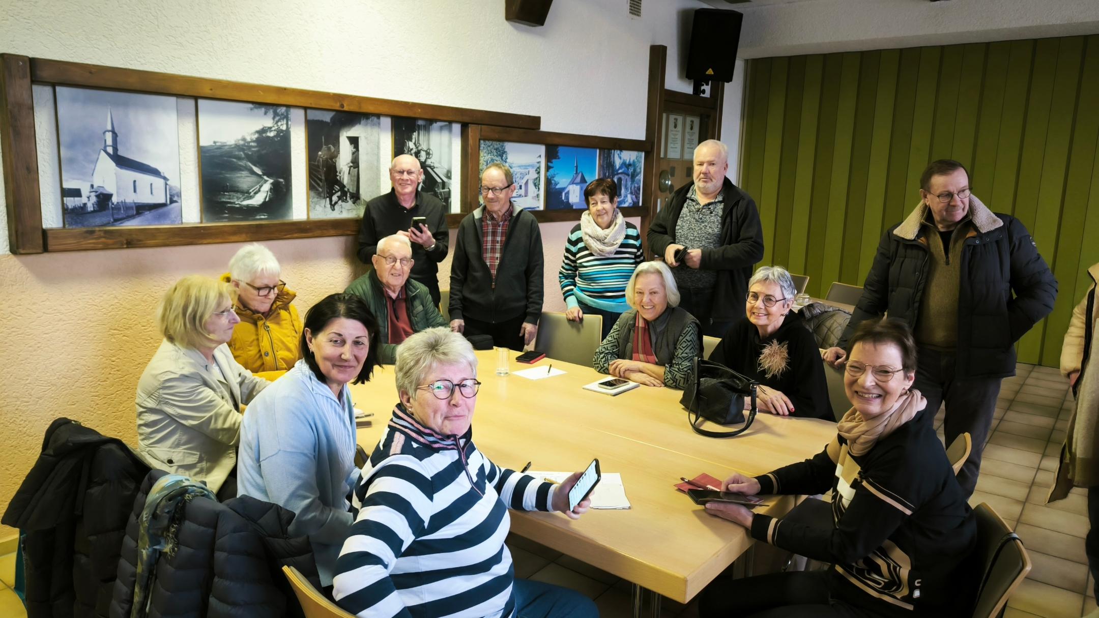 Eine Gruppe von Menschen sitzt und steht gemeinsam in einem Raum um einen Tisch, viele mit Smartphones in der Hand, als Teil einer Runde zum Austausch oder Lernen.