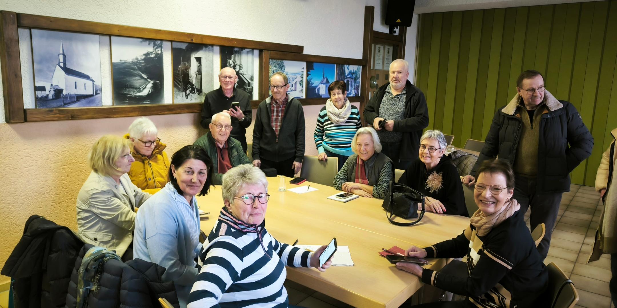 Eine Gruppe von Menschen sitzt und steht gemeinsam in einem Raum um einen Tisch, viele mit Smartphones in der Hand, als Teil einer Runde zum Austausch oder Lernen.