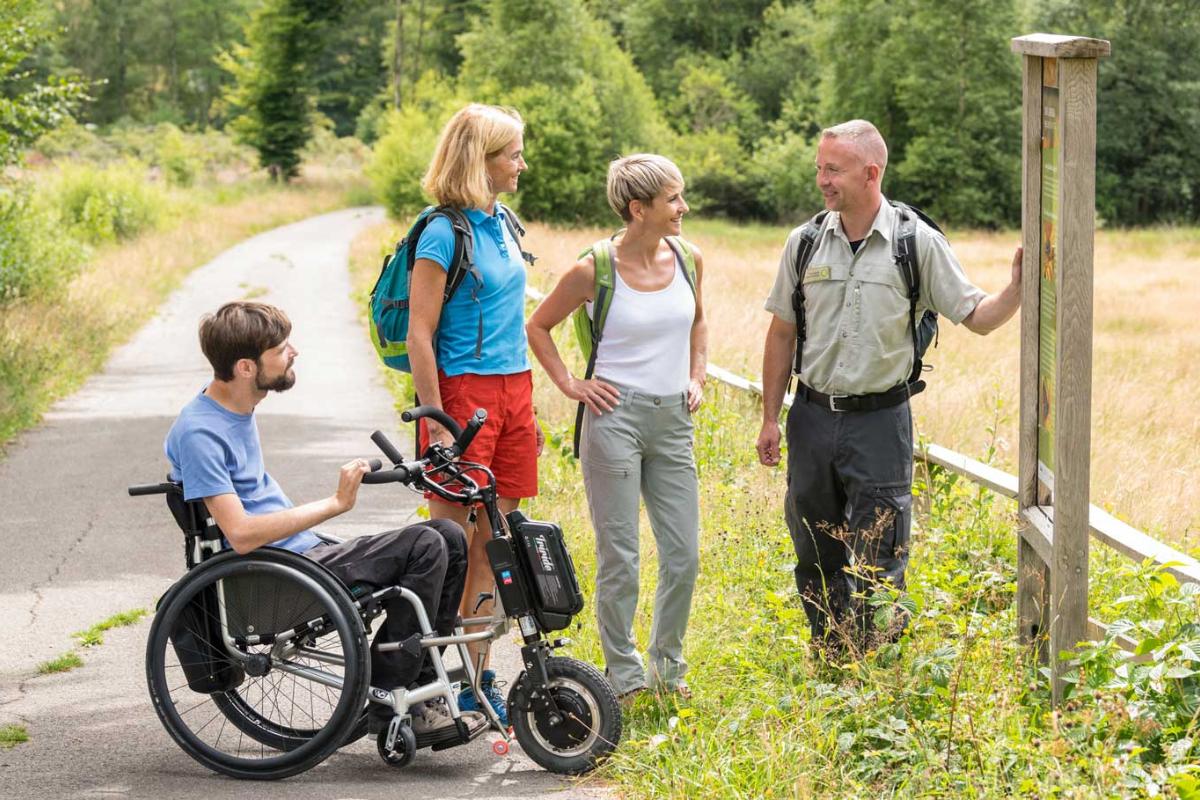 Vier Personen auf einem Naturweg, eine im Rollstuhl, im Gespräch an einer Infotafel