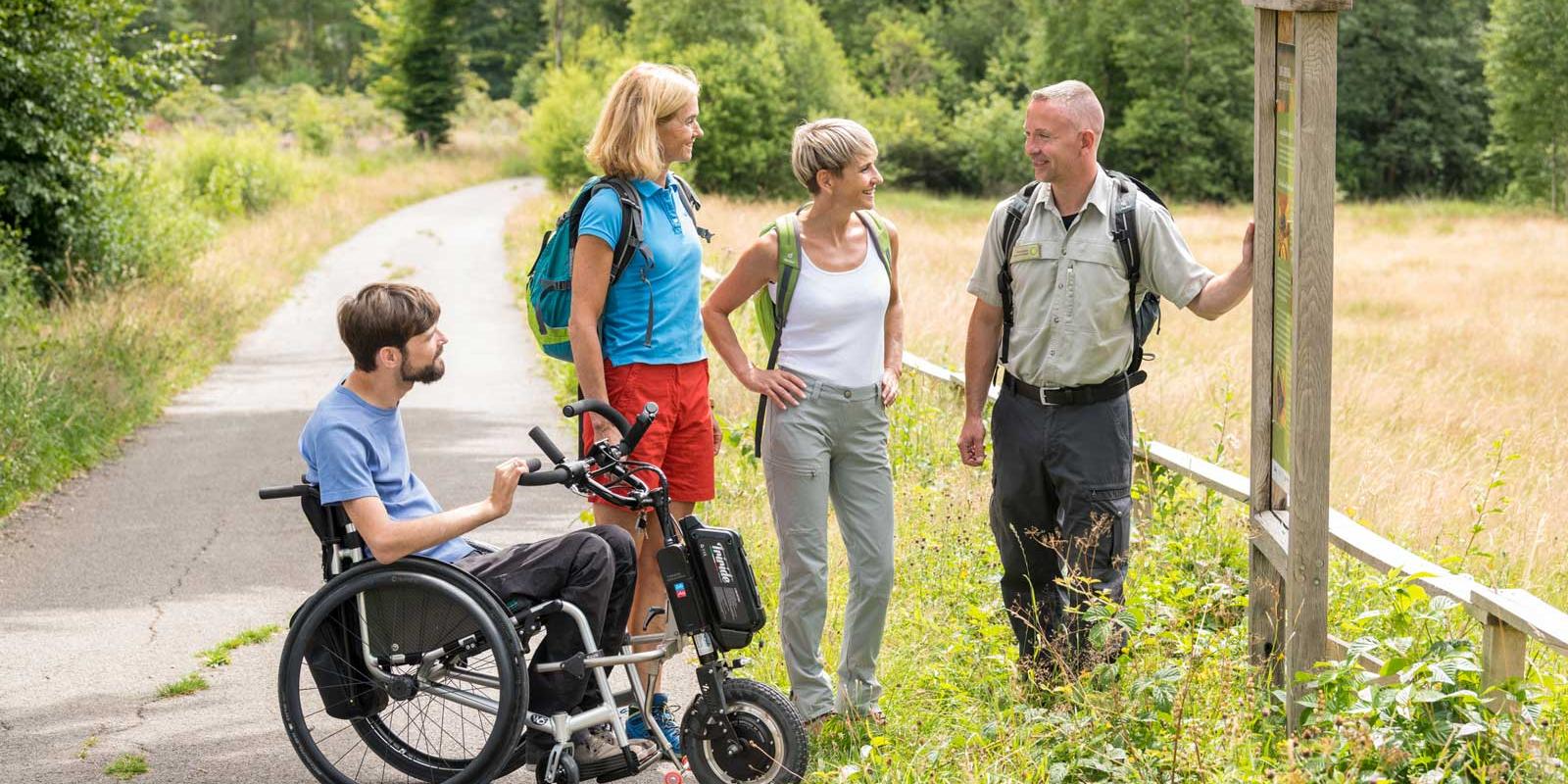 Vier Personen auf einem Naturweg, eine im Rollstuhl, im Gespräch an einer Infotafel
