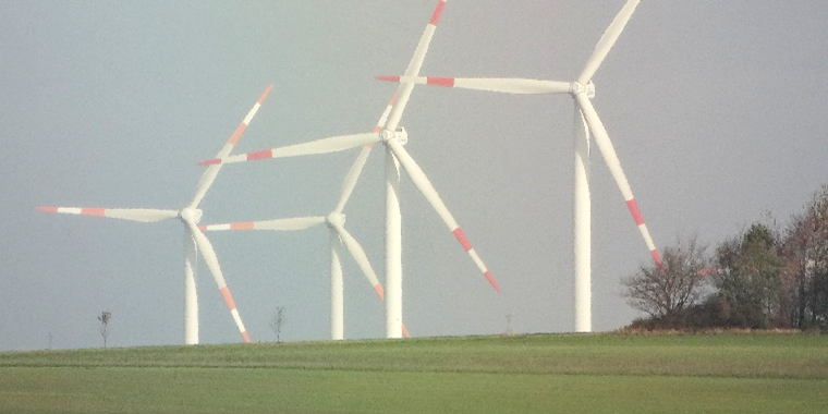 Das Foto zeigt vier große Windräder auf einem grünen Feld, eingerahmt von Bäumen im Vordergrund und einem bewölkten Himmel im Hintergrund.