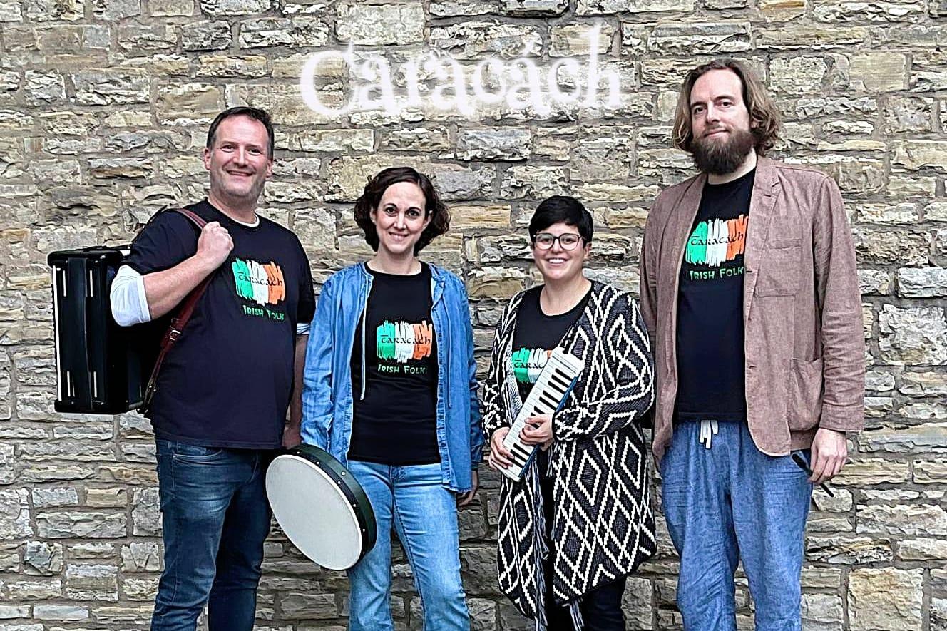 Copilot said:  Vier Personen der Irish-Folk-Band stehen mit Instrumenten vor einer Natursteinmauer mit dem Schriftzug „Caracách“.