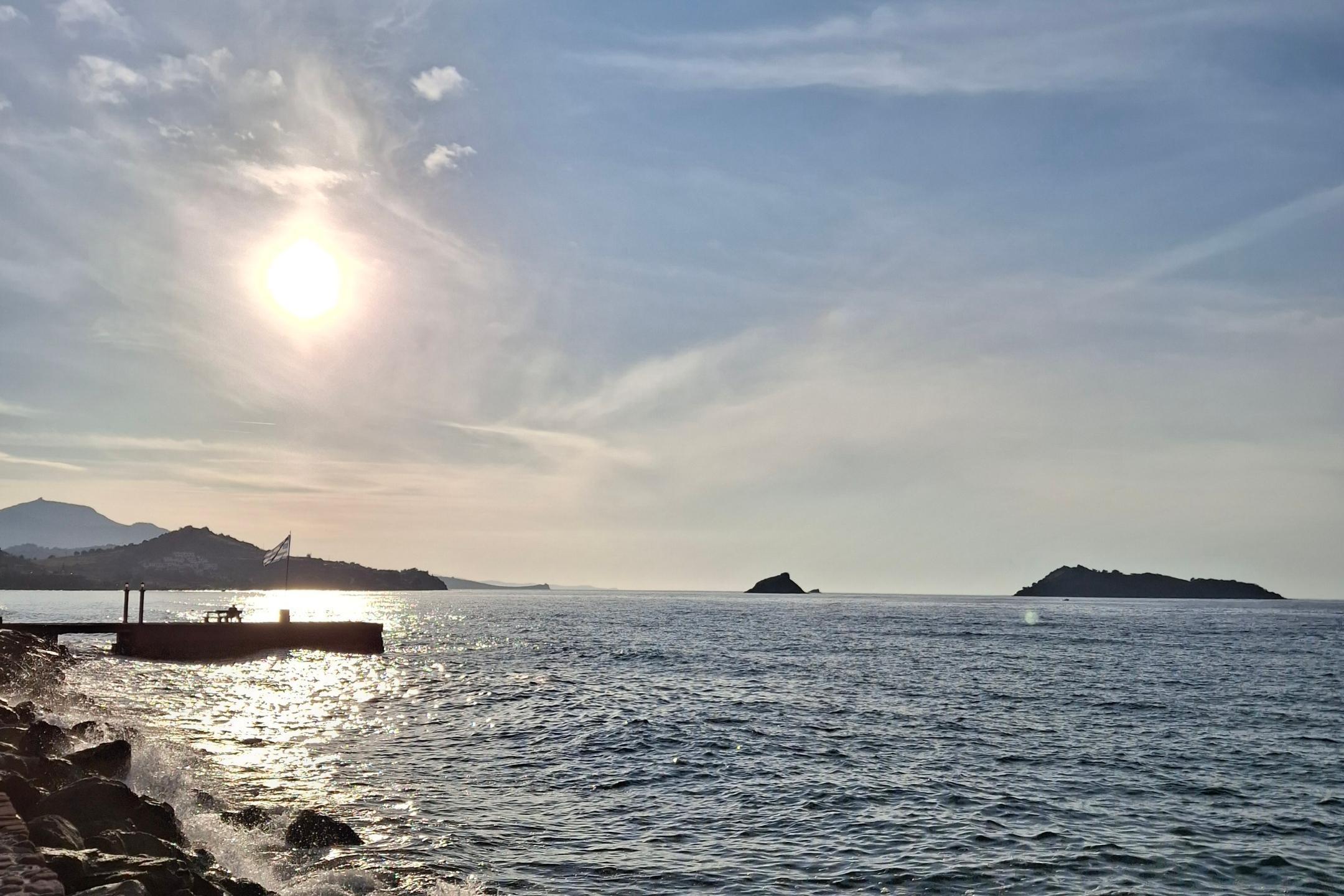 Das Bild zeigt eine sonnige Küstenlandschaft mit ruhiger See, Wellen an einer Steinmole und einer strahlenden Sonne über einem leicht bewölkten Himmel.