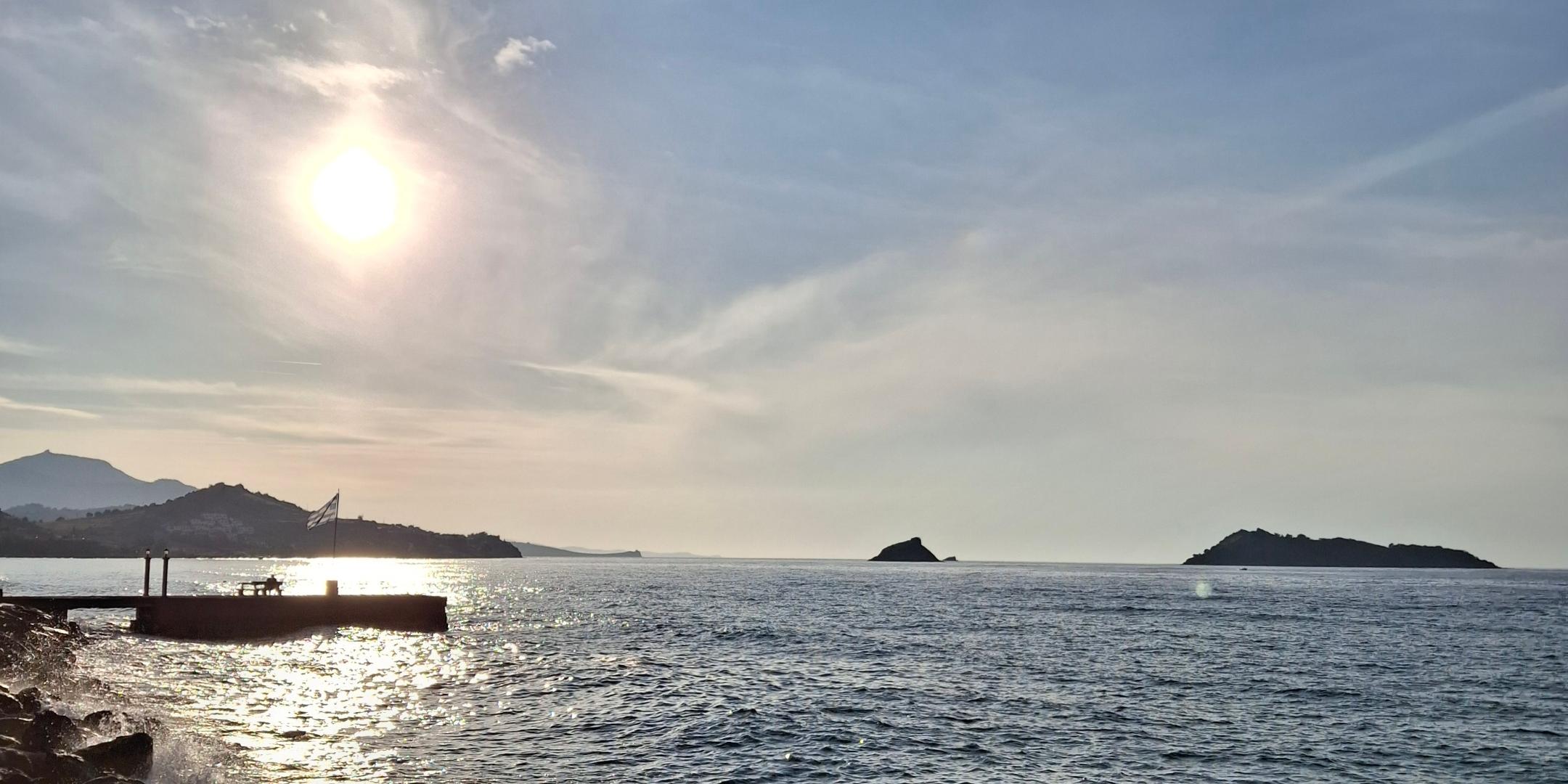 Das Bild zeigt eine sonnige Küstenlandschaft mit ruhiger See, Wellen an einer Steinmole und einer strahlenden Sonne über einem leicht bewölkten Himmel.