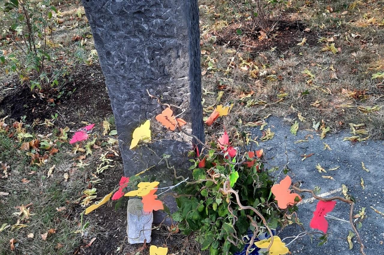 Ein hoher Gedenkstein mit einer Schmetterlingsskulptur oben steht auf einem Friedhof. Davor wächst ein Strauch, an dem bunte Papierschmetterlinge hängen.