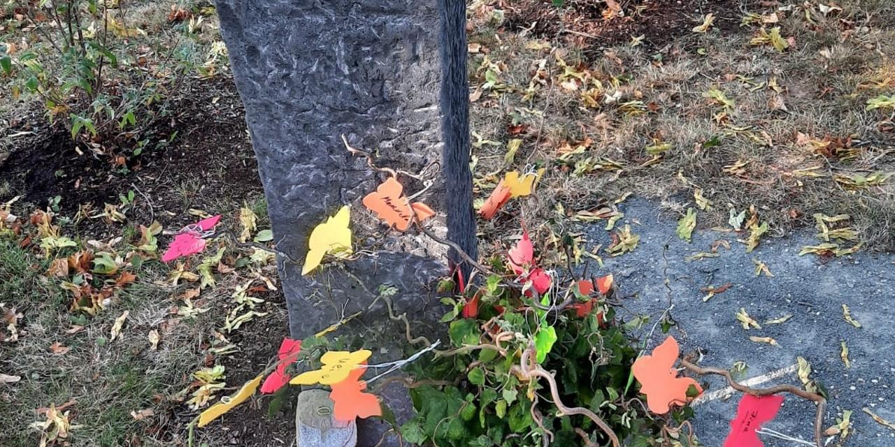Ein hoher Gedenkstein mit einer Schmetterlingsskulptur oben steht auf einem Friedhof. Davor wächst ein Strauch, an dem bunte Papierschmetterlinge hängen.