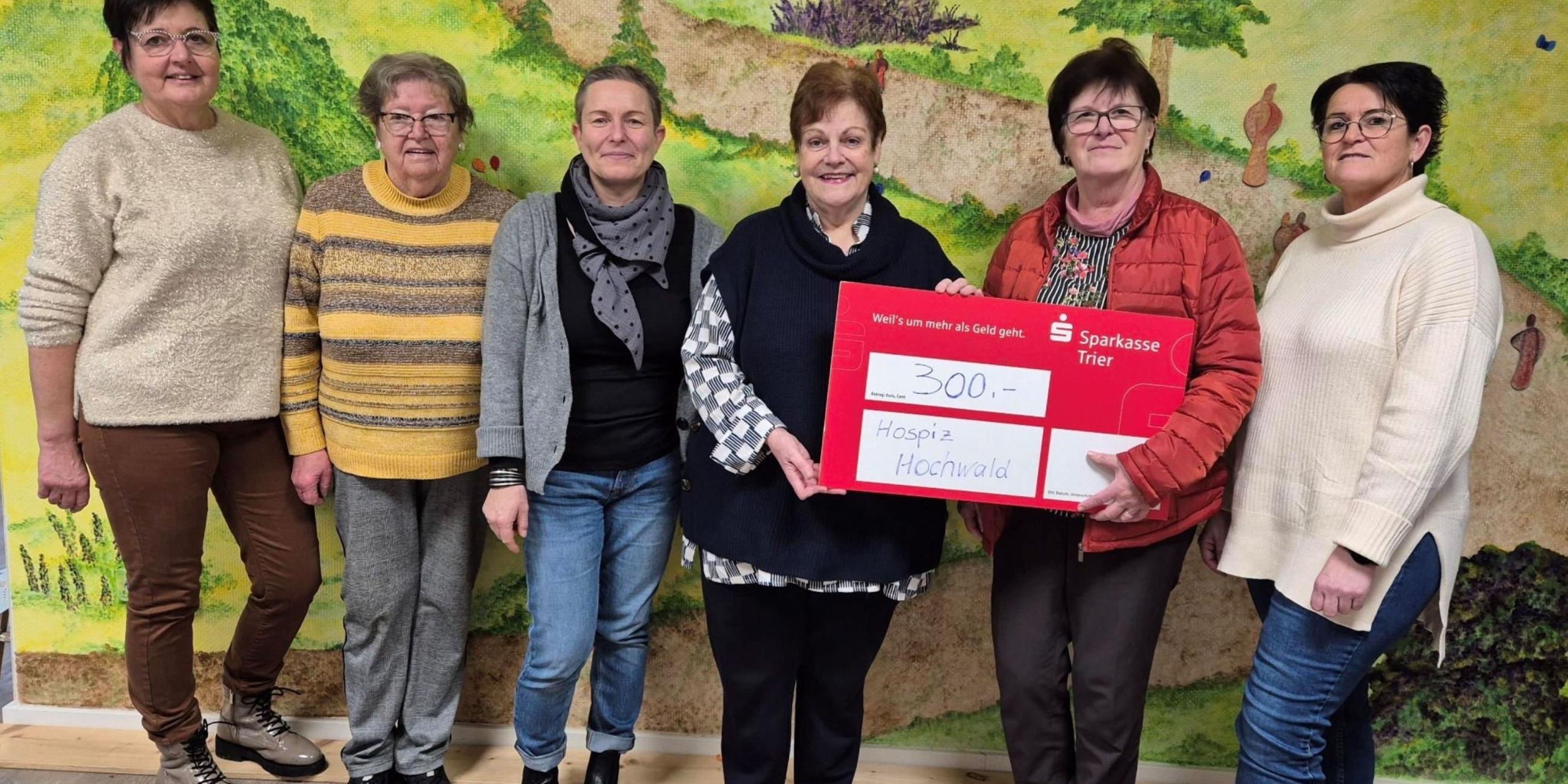 Sechs Personen stehen vor einer Wand mit einem bunten Landschaftsgemälde und halten gemeinsam einen großen roten Spendenscheck über 300 Euro für das „Hospiz Hochwald (c) Ambulantes Hospiz Hochwald Sechs Personen stehen vor einer Wand mit einem bunten Landschaftsgemälde und halten gemeinsam einen großen roten Spendenscheck über 300 Euro für das „Hospiz Hochwald
