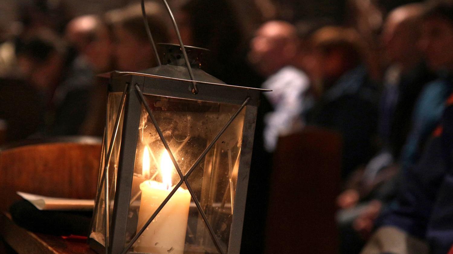 Das Foto zeigt eine Laterne mit einer brennenden Kerze, die auf einer Kirchenbank steht, während im Hintergrund Menschen in einer Kirche sitzen.