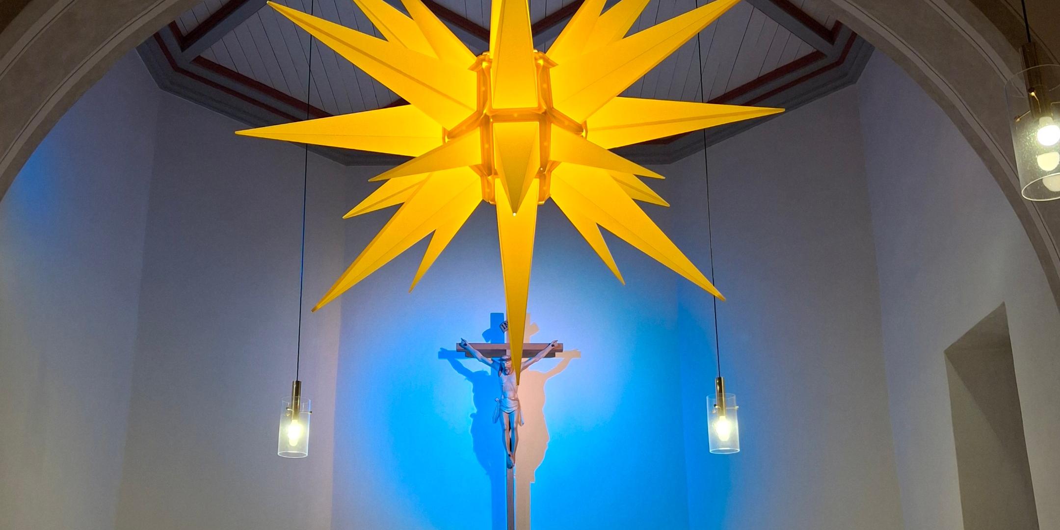 Das Bild zeigt einen hell erleuchteten Kirchenaltar mit einer großen gelben Weihnachtsstern-Laterne im Vordergrund, Kerzen und Blumen auf dem Altar sowie einem goldenen Tabernakel, während ein blau angestrahltes Kreuz im Hintergrund hängt.