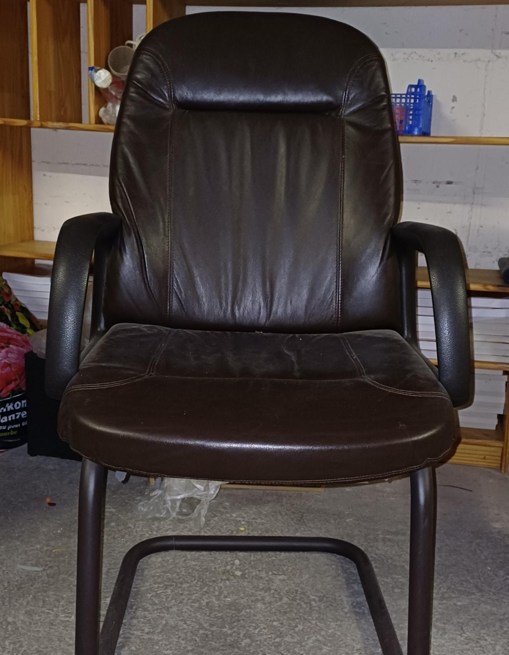 Der Stuhl ist aus braunem Leder gefertigt, hat Armlehnen und steht auf einem stabilen Metallgestell.