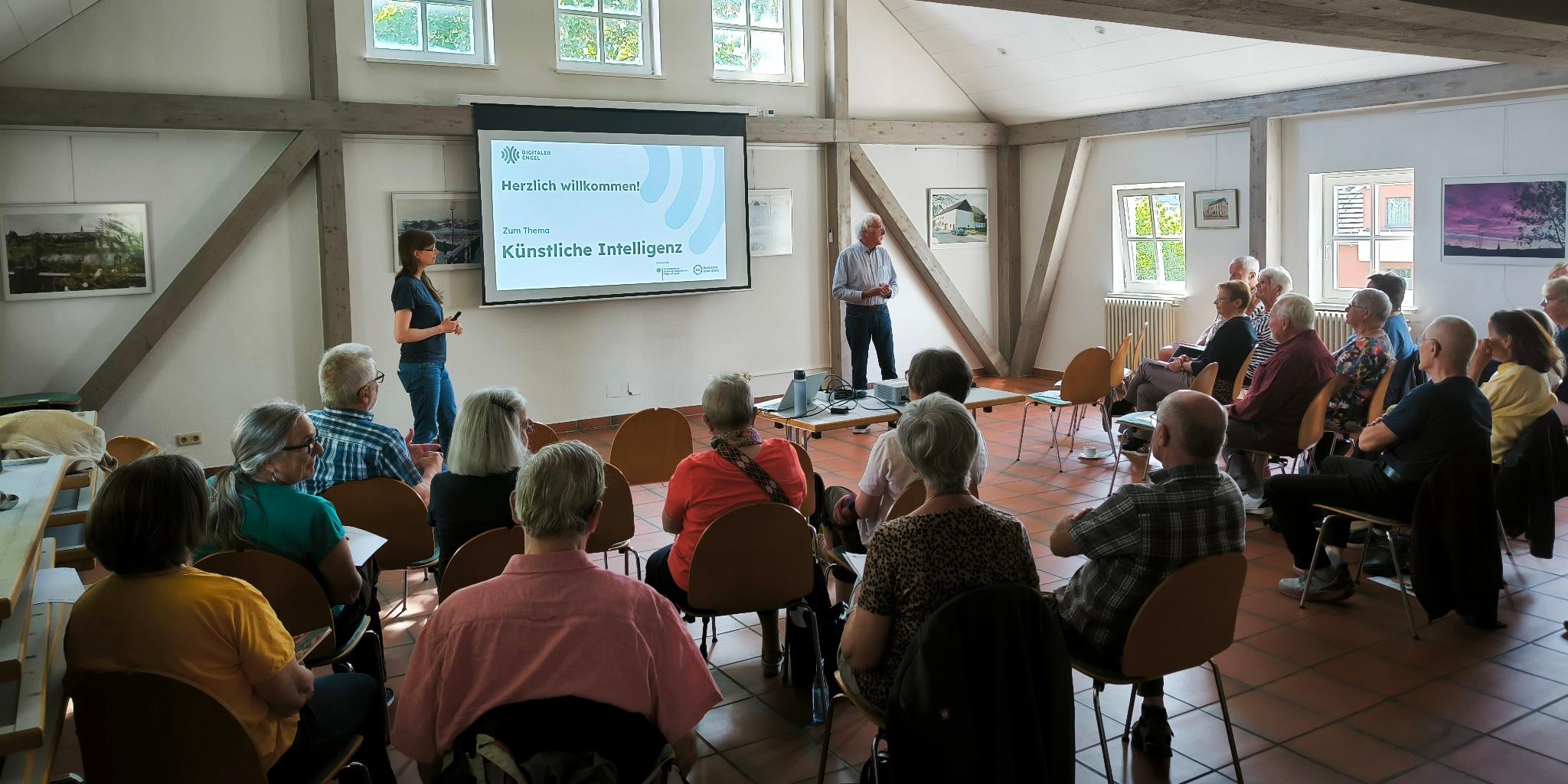 Teilnehmende verfolgen in einem hellen Saal mit Holzbalken aufmerksam einen Vortrag der Digitalen Engel zum Thema Künstliche Intelligenz.