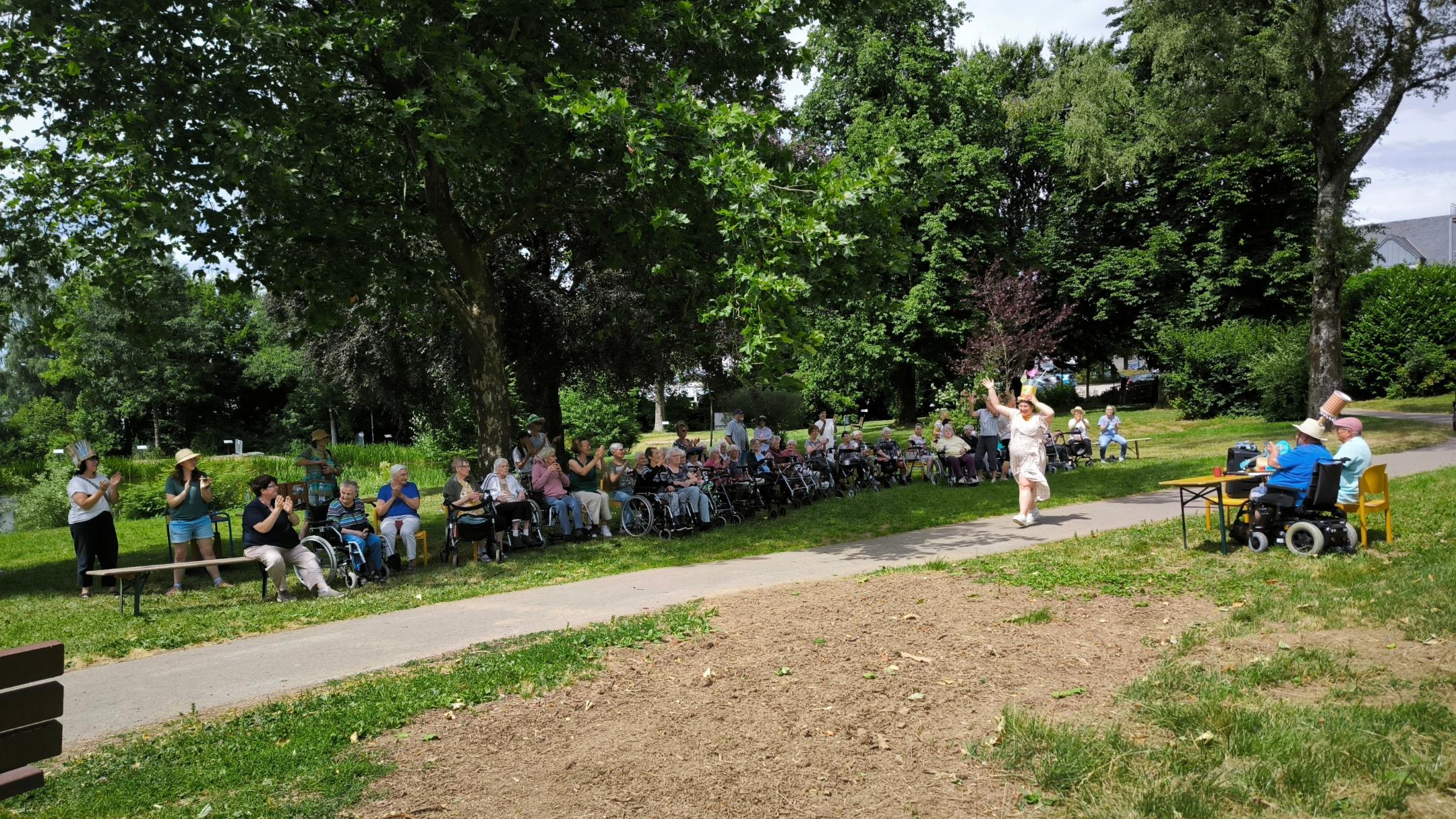 Eine Menschengruppe bei der Veranstaltung „Verrückte Hüte“ im Stadtpark unter großen Bäumen.