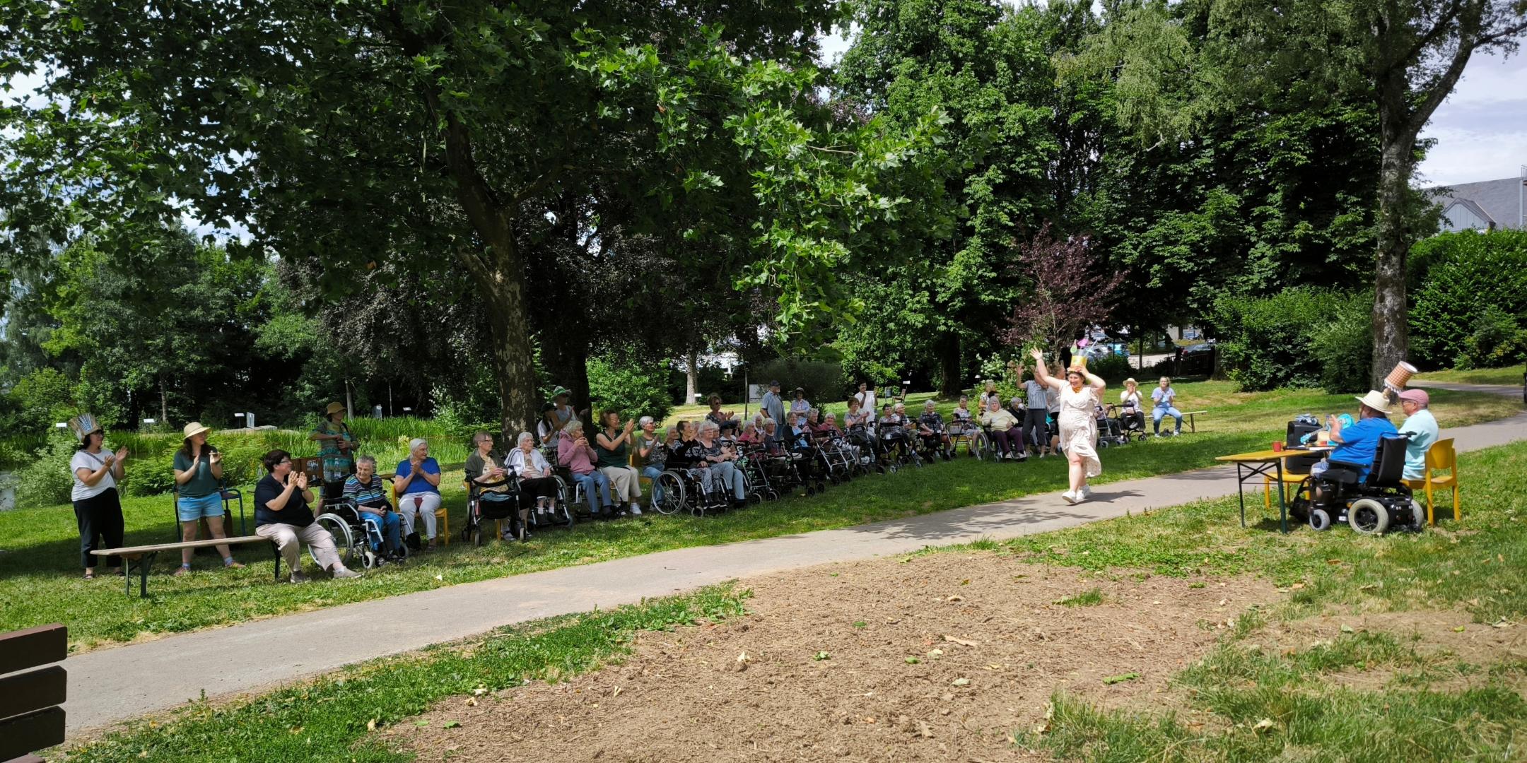 Eine Menschengruppe bei der Veranstaltung „Verrückte Hüte“ im Stadtpark unter großen Bäumen.