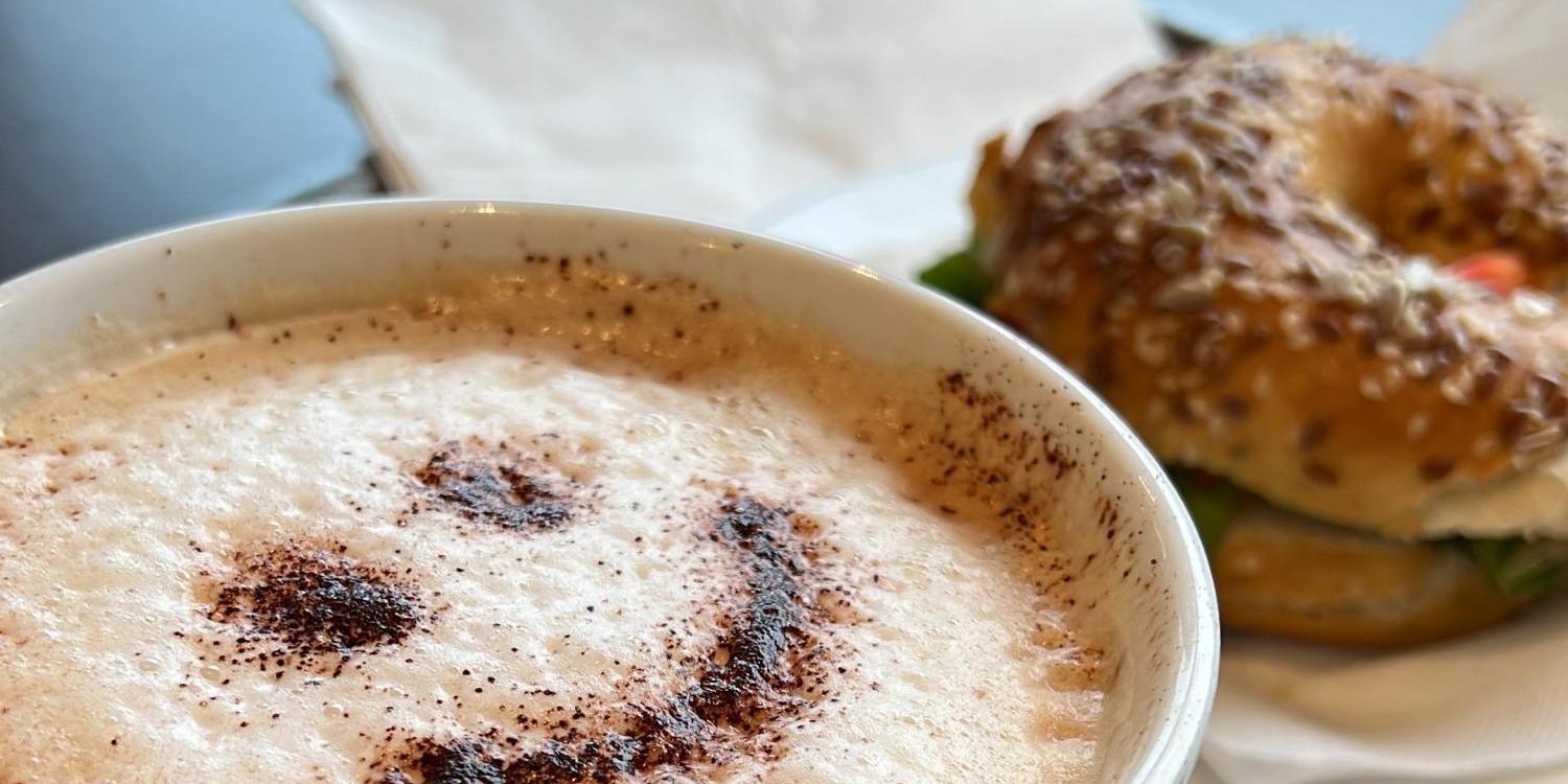 Eine Tasse Cappuccino mit Smiley aus Kakao steht neben einem Bagel und einem Stück Kuchen.