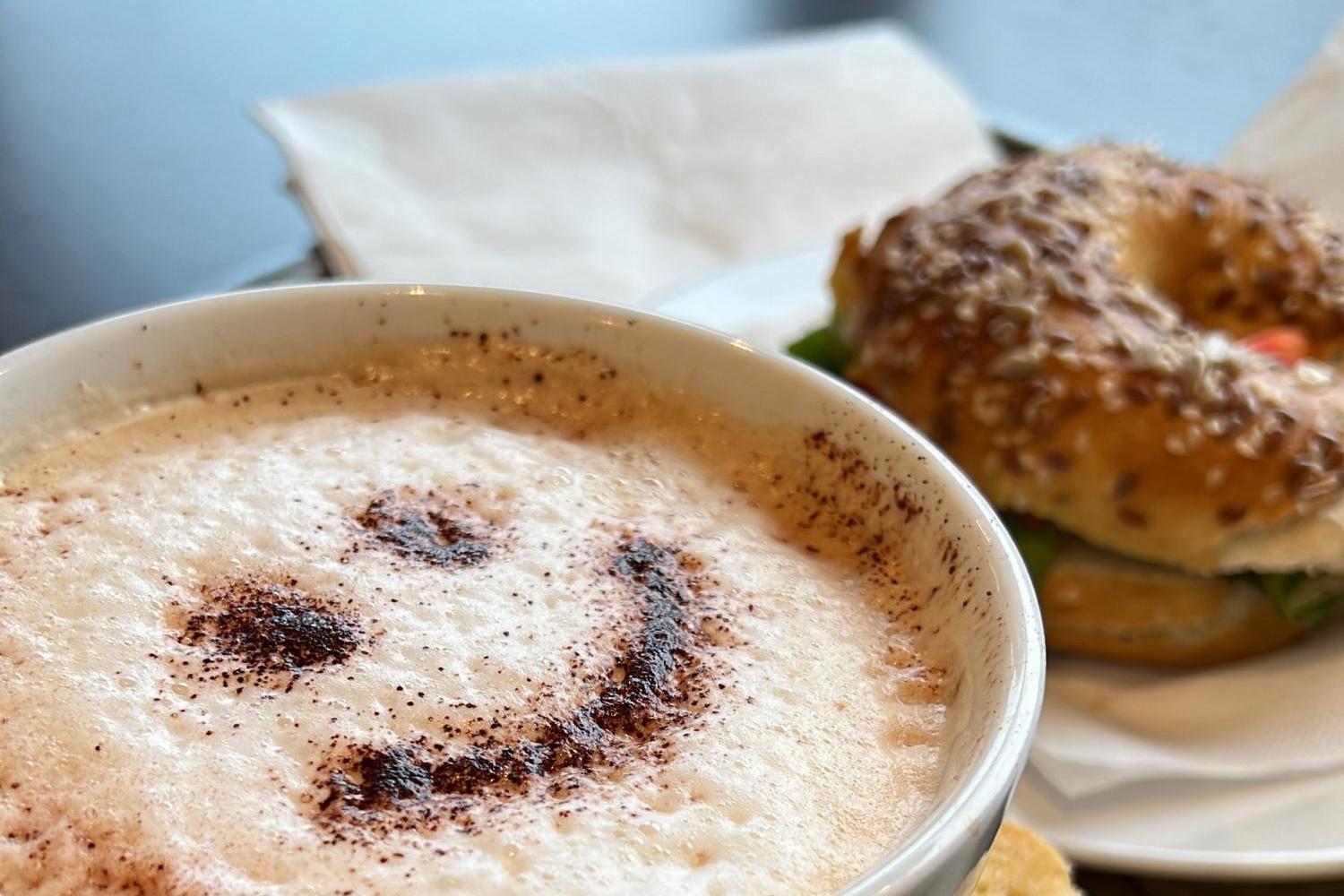Eine Tasse Cappuccino mit Smiley aus Kakao steht neben einem Bagel und einem Stück Kuchen.