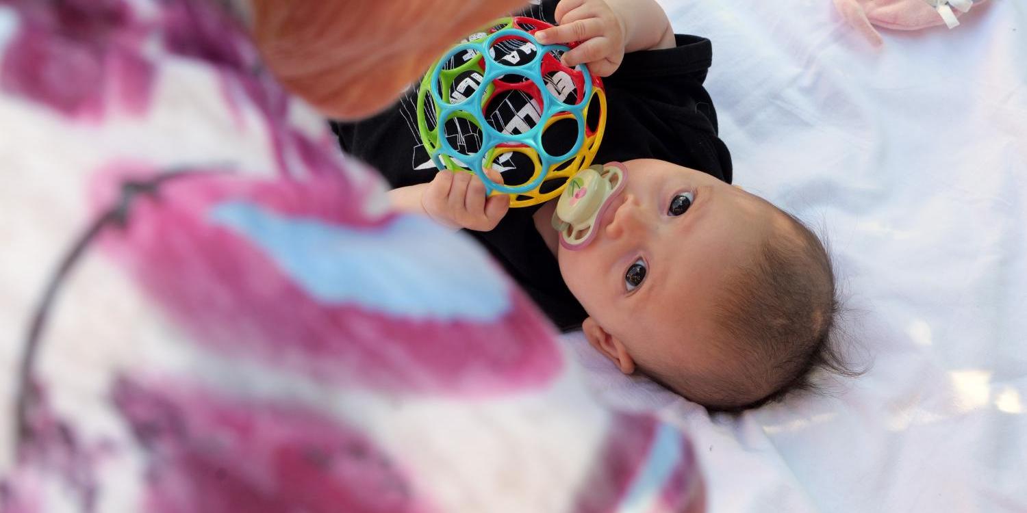 Eine ältere Person beugt sich über ein kleines Kind, das auf einer Decke liegt und mit einem bunten Spielball spielt.