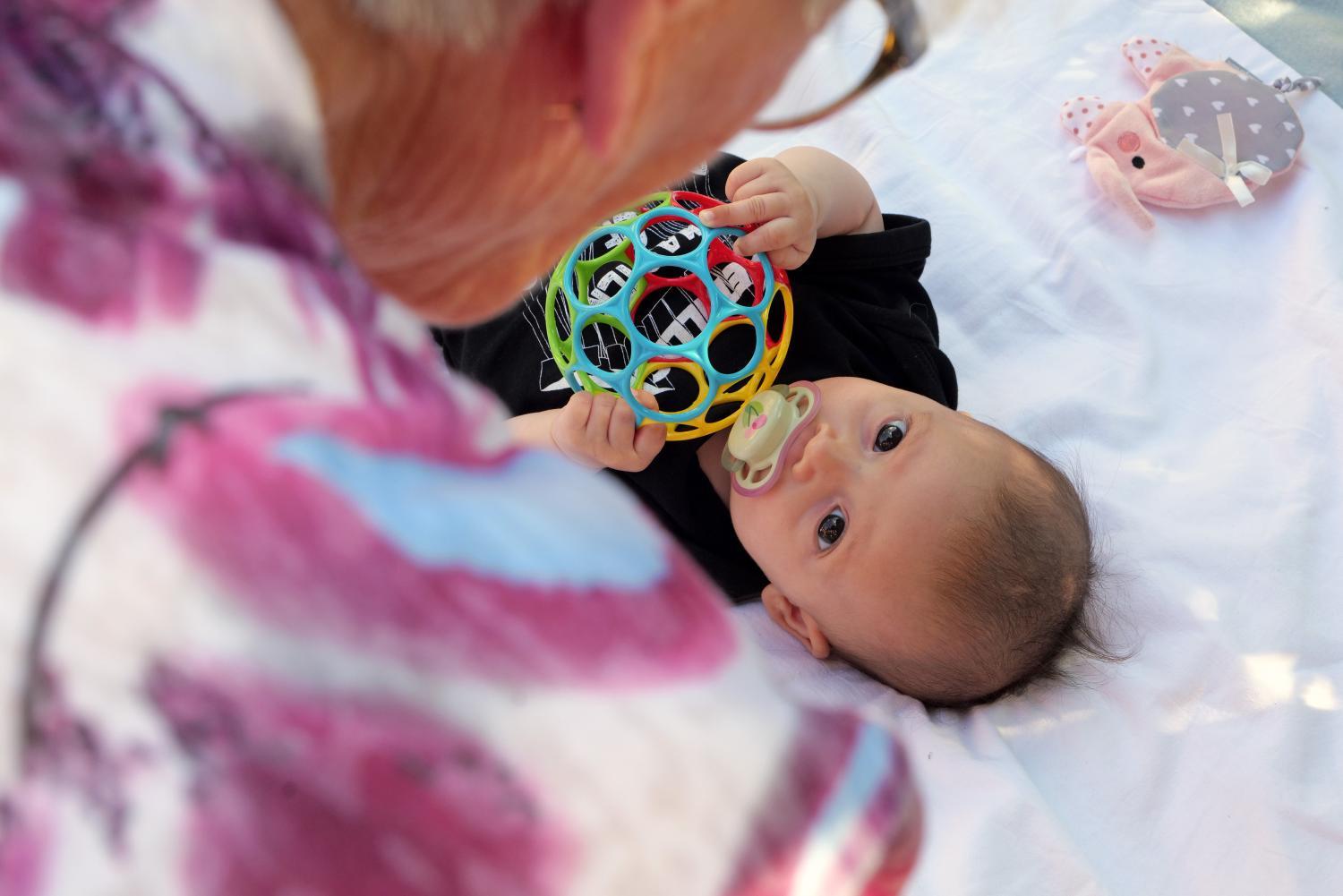 Eine ältere Person beugt sich über ein kleines Kind, das auf einer Decke liegt und mit einem bunten Spielball spielt.