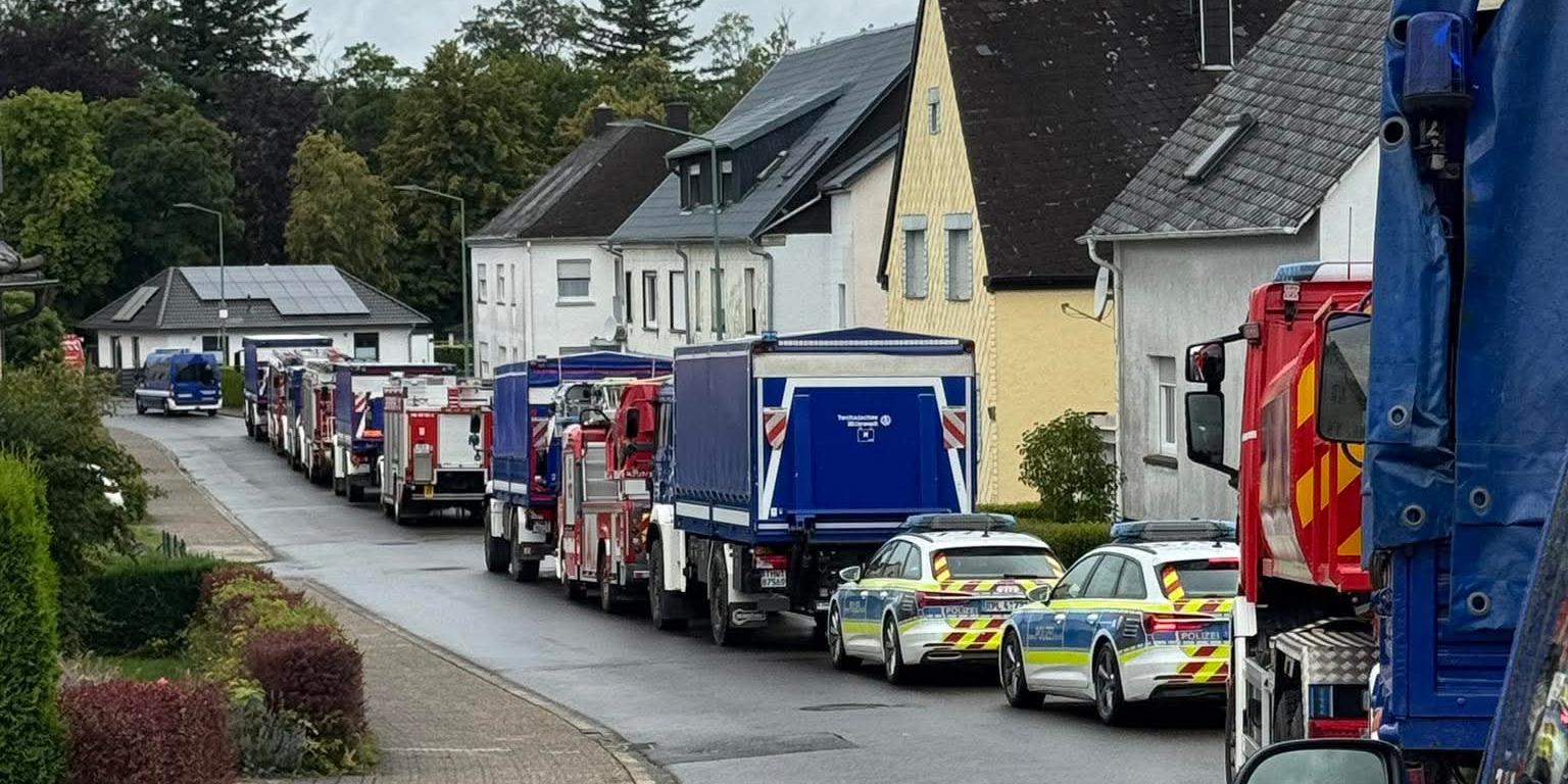 Nasse Wohnstraße mit einer langen Reihe von Feuerwehrfahrzeugen und Polizeiautos vor Wohnhäusern unter grauem Himmel.