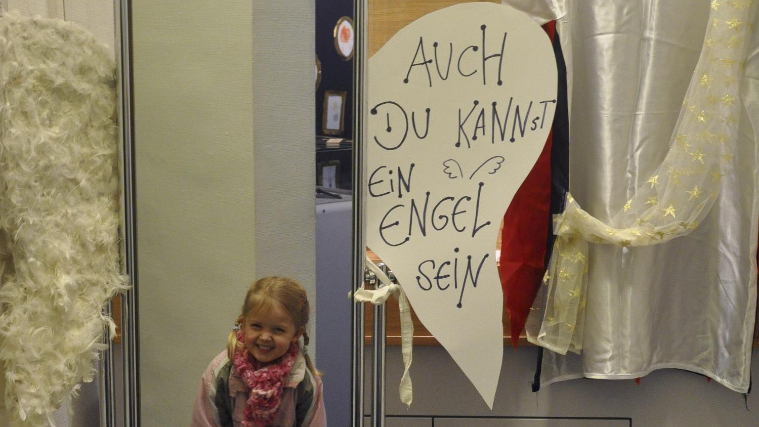 Ein Kind steht vor einem großen Spiegel, daneben hängen weiße Engelsflügel und ein weißes Gewand, mit einem Schild: „Auch du kannst ein Engel sein“.