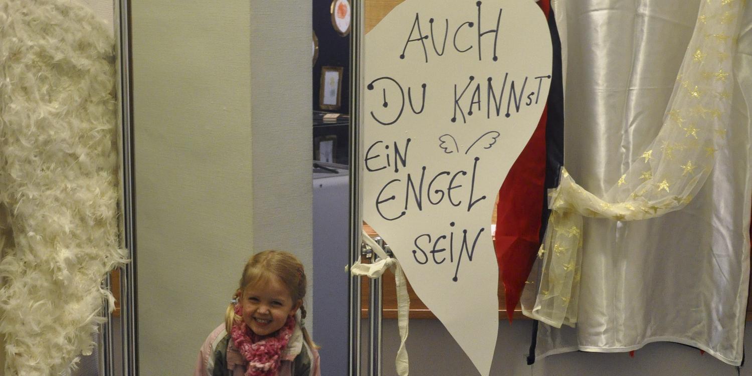 Ein Kind steht vor einem großen Spiegel, daneben hängen weiße Engelsflügel und ein weißes Gewand, mit einem Schild: „Auch du kannst ein Engel sein“.