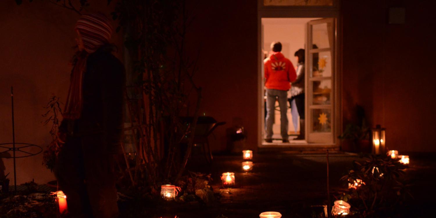 Das Foto zeigt einen dunklen Gartenweg, der mit vielen leuchtenden Kerzen in Gläsern gesäumt ist und zu einer offenen Tür führt, hinter der Menschen in einem warm erleuchteten Raum stehen.