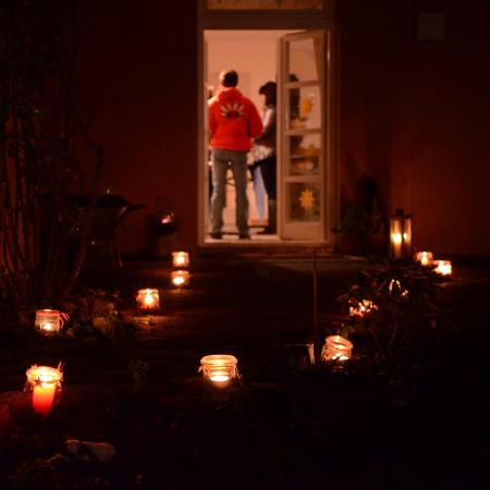 Das Foto zeigt einen dunklen Gartenweg, der mit vielen leuchtenden Kerzen in Gläsern gesäumt ist und zu einer offenen Tür führt, hinter der Menschen in einem warm erleuchteten Raum stehen.