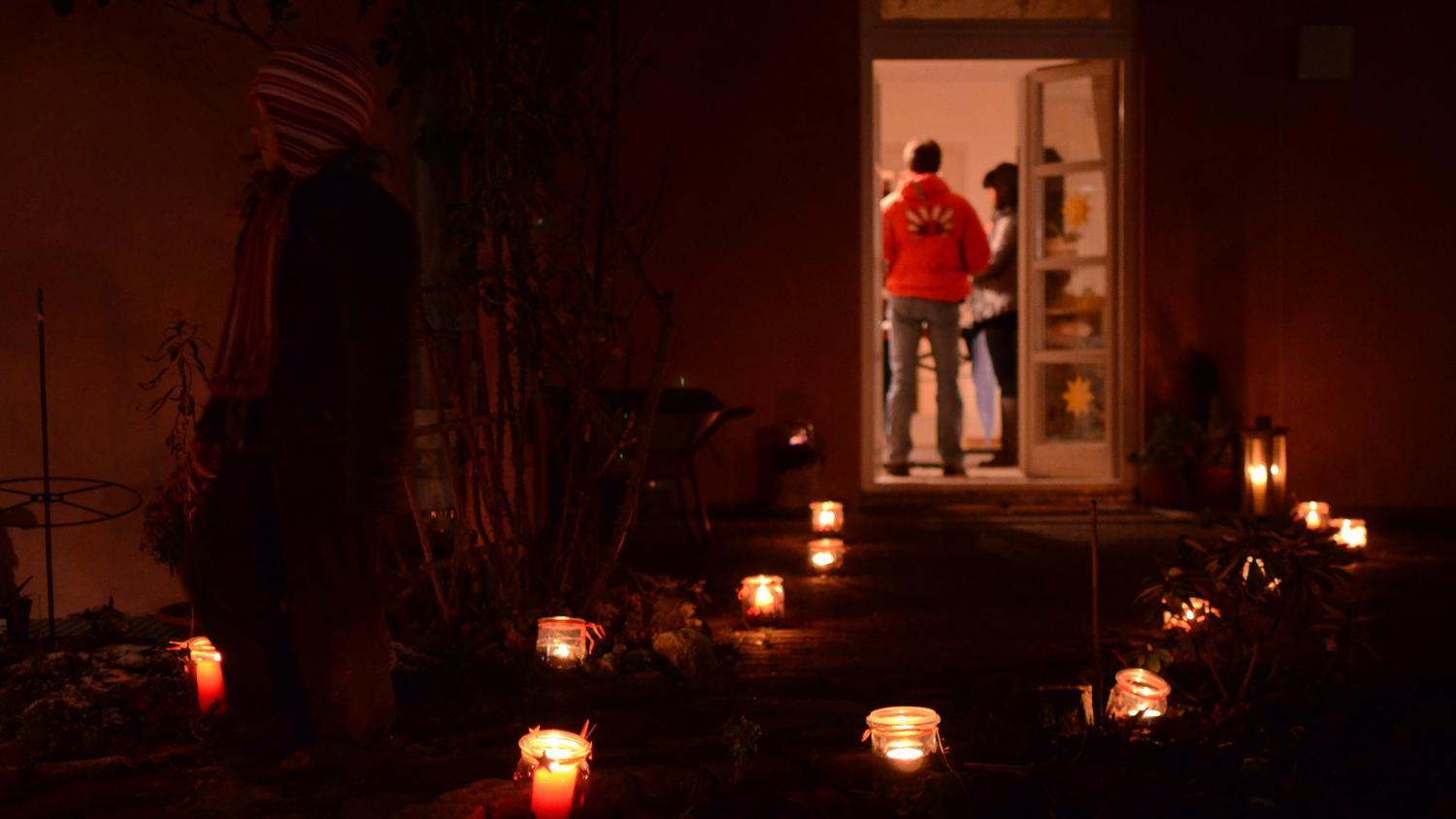 Das Foto zeigt einen dunklen Gartenweg, der mit vielen leuchtenden Kerzen in Gläsern gesäumt ist und zu einer offenen Tür führt, hinter der Menschen in einem warm erleuchteten Raum stehen.