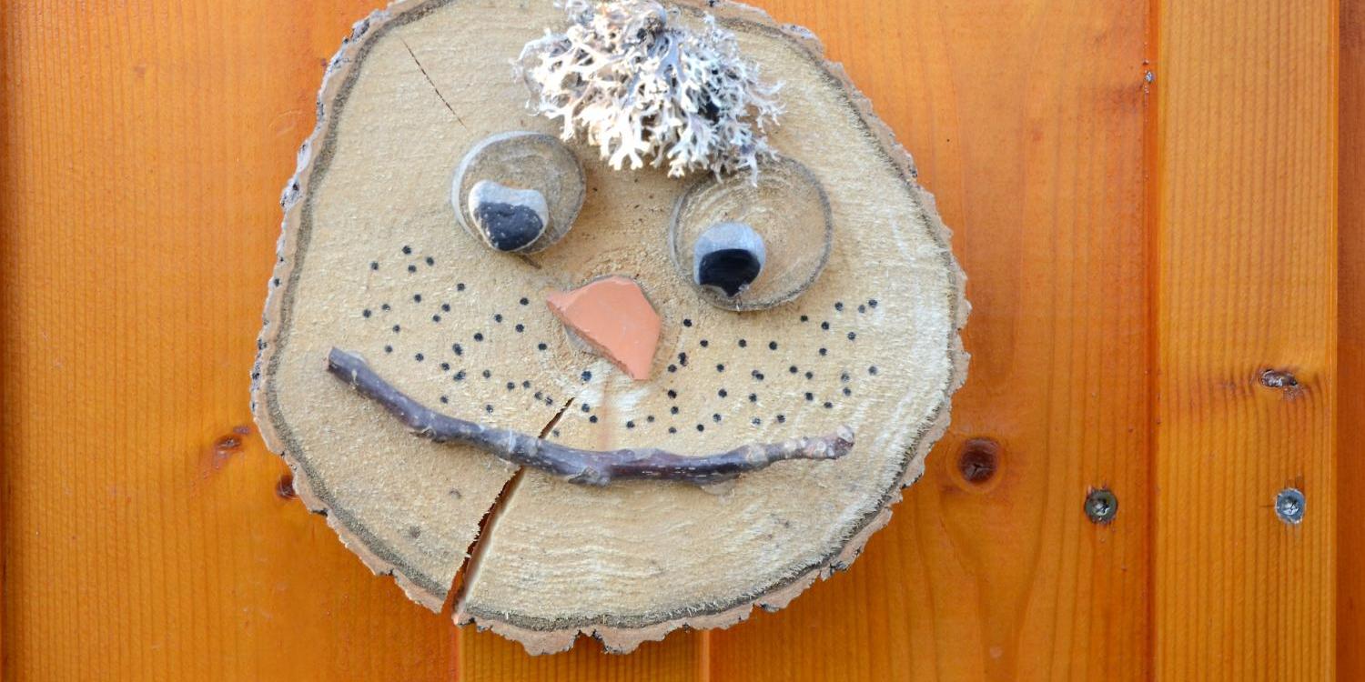 Holzscheibe mit Gesicht aus Naturmaterialien an Holzwand befestigt.
