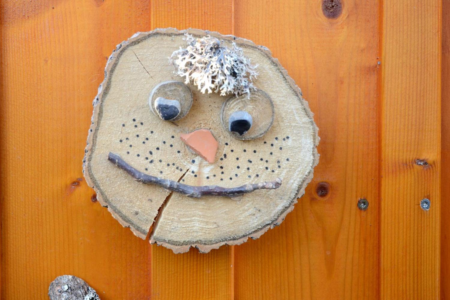 Holzscheibe mit Gesicht aus Naturmaterialien an Holzwand befestigt.