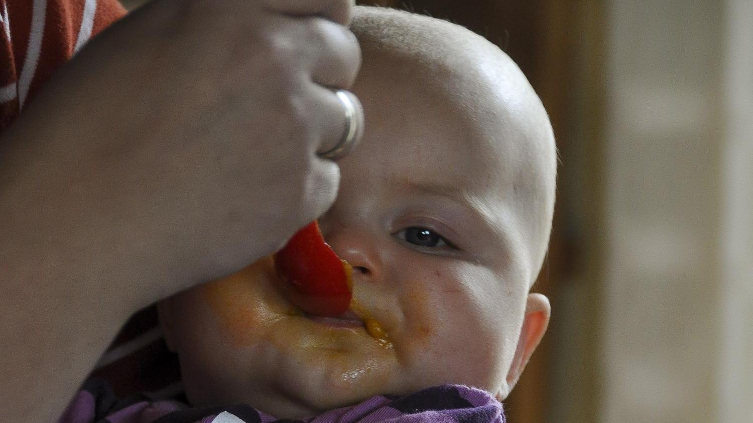 Das Bild zeigt eine Person, die ein kleines Kind in den Armen hält, während vor ihnen auf dem Tisch eine Schüssel mit Speiseresten steht.