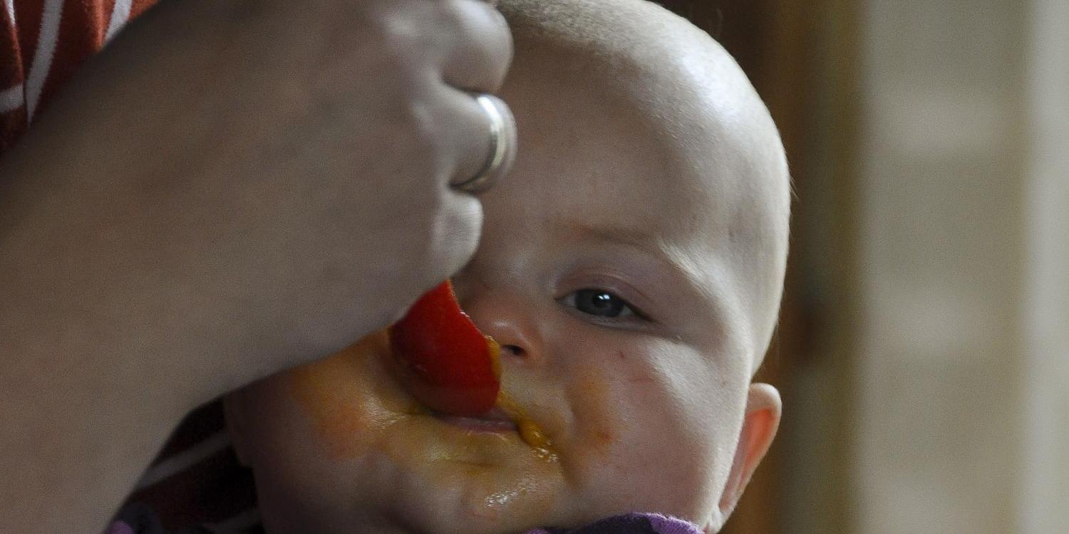 Das Bild zeigt eine Person, die ein kleines Kind in den Armen hält, während vor ihnen auf dem Tisch eine Schüssel mit Speiseresten steht.