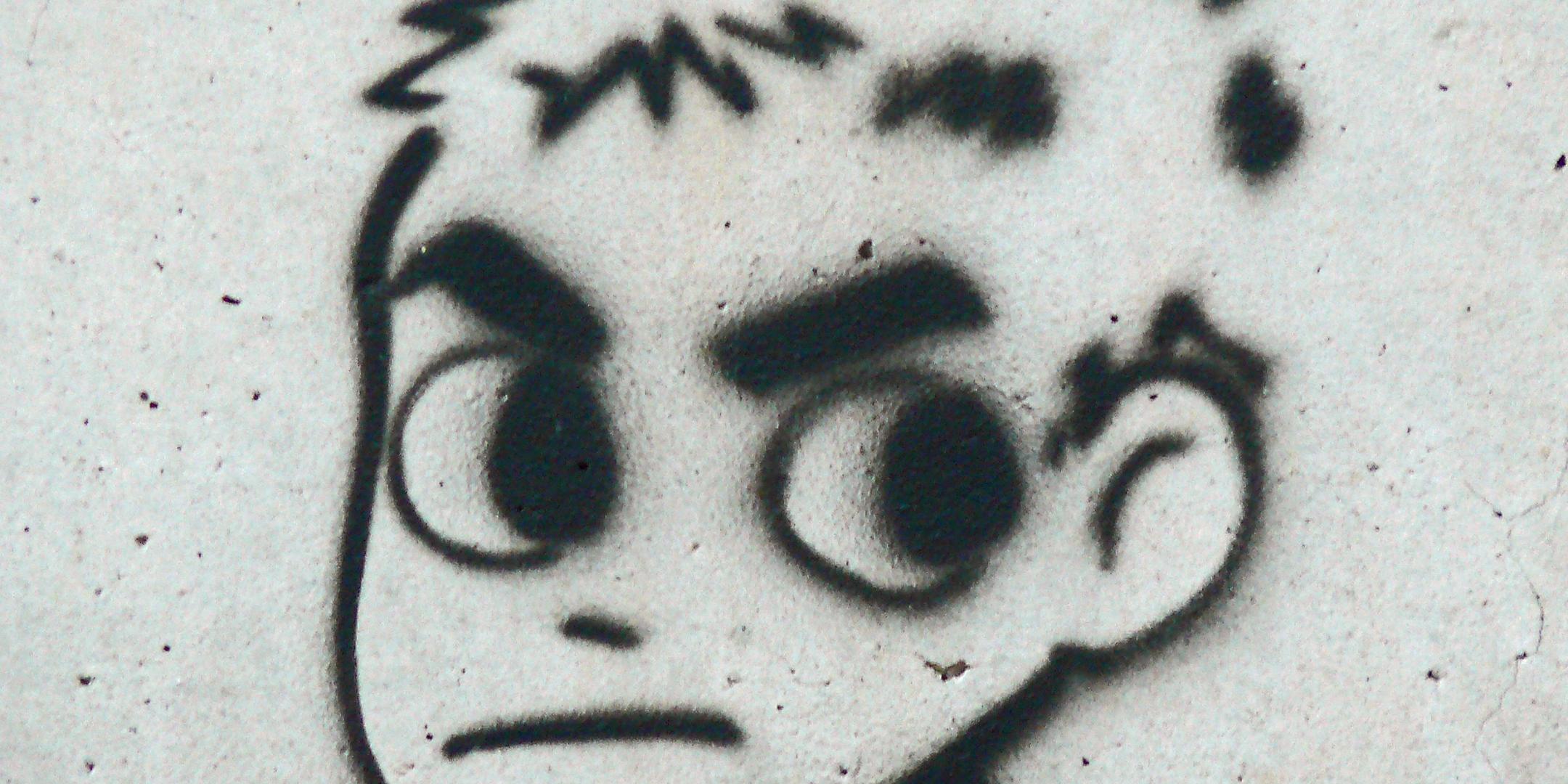 Das Bild zeigt eine Wand mit einem Graffiti, das in schwarzer Farbe geschwungene Linien und Formen darstellt, die wie Teile eines stilisierten Gesichts wirken.