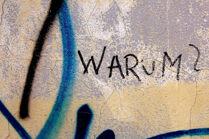 Auf einer verwitterten Wand steht in schwarzer Schrift das Wort „WARUM?“, umgeben von blauer Graffiti-Farbe.