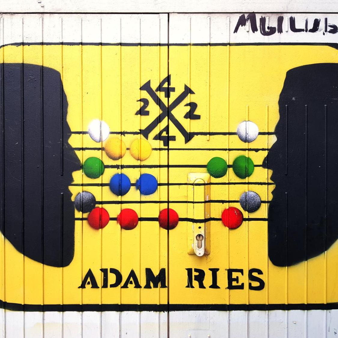 Gelbes Bild mit zwei schwarzen Kopf-Silhouetten und einem bunten Abakus, darunter der Schriftzug ‚Adam Ries‘.