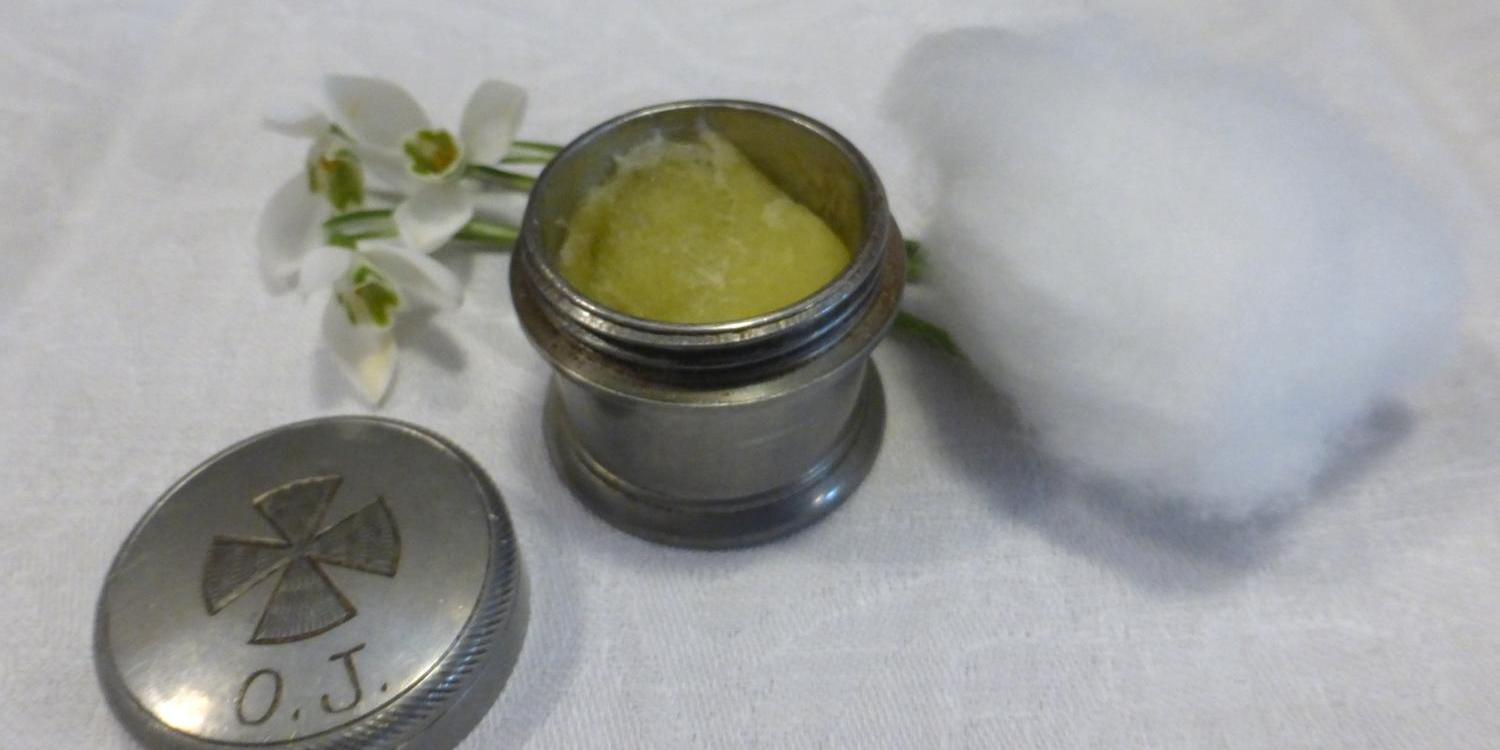 Das Foto zeigt ein kleines silbernes Gefäß mit geöffnetem Deckel, das gelbliche Salbe enthält, daneben liegen ein Stück Watte und einige weiße Blüten auf einem weißen Tuch.