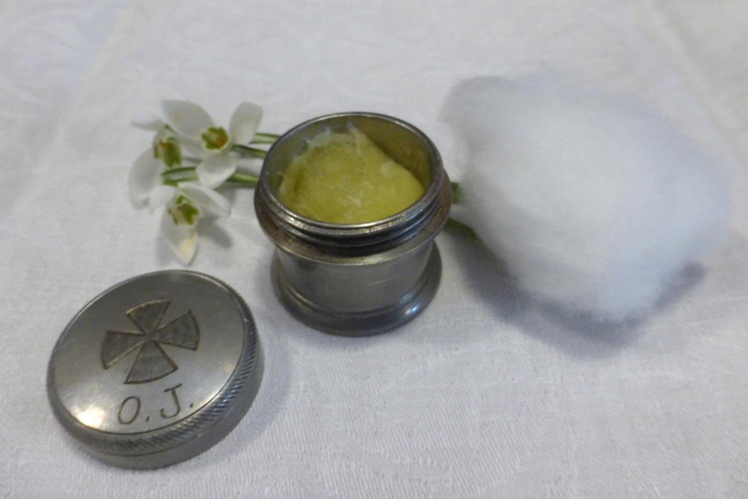 Das Foto zeigt ein kleines silbernes Gefäß mit geöffnetem Deckel, das gelbliche Salbe enthält, daneben liegen ein Stück Watte und einige weiße Blüten auf einem weißen Tuch.