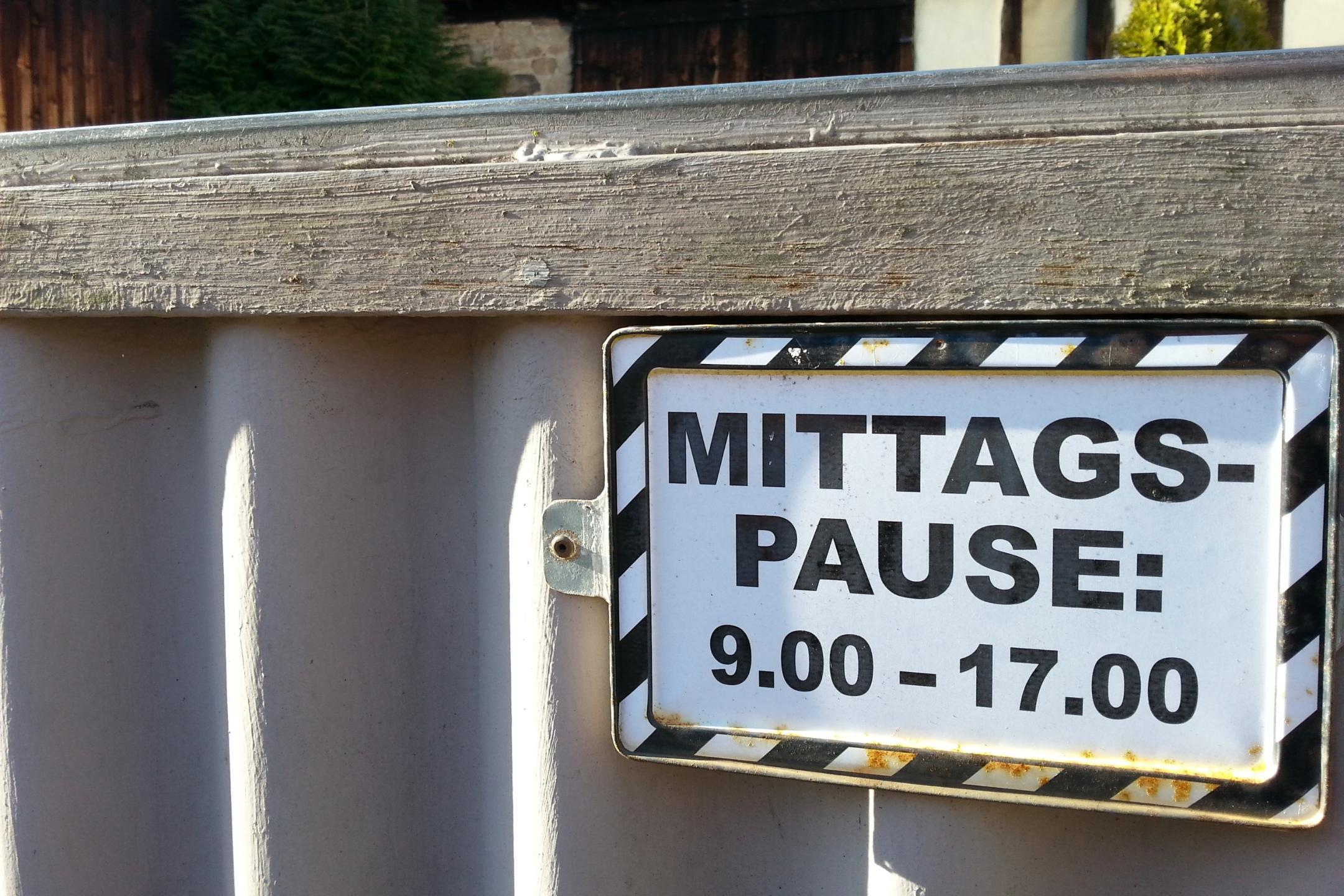 Das Foto zeigt ein Schild mit der Aufschrift „MITTAGSPAUSE: 9.00 – 17.00“, befestigt an einem Metallgeländer vor einem Gebäude mit Holz- und Steinfassade.