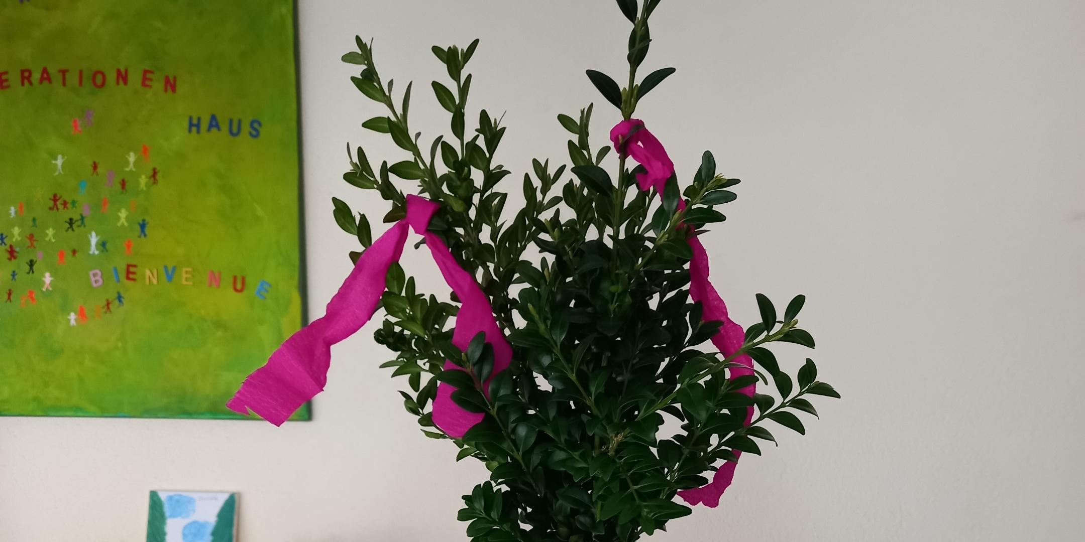 Ein kleiner geschmückter Strauch in einem weißen Topf steht auf einem Tisch mit einer grünen Filzunterlage.