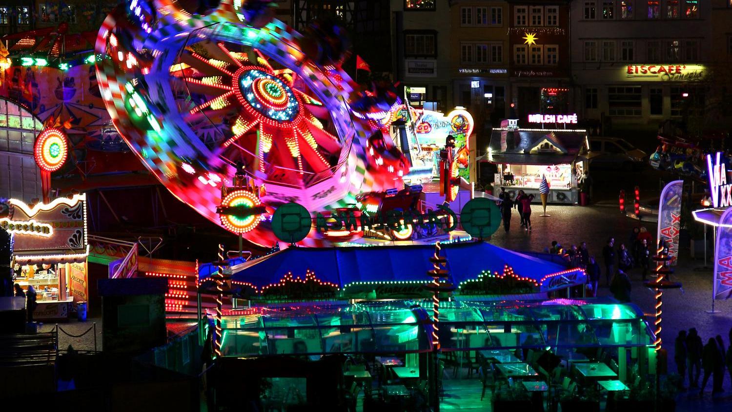 Bunter Jahrmarkt bei Nacht mit hell erleuchtetem Fahrgeschäft und leeren Tischen im Vordergrund.