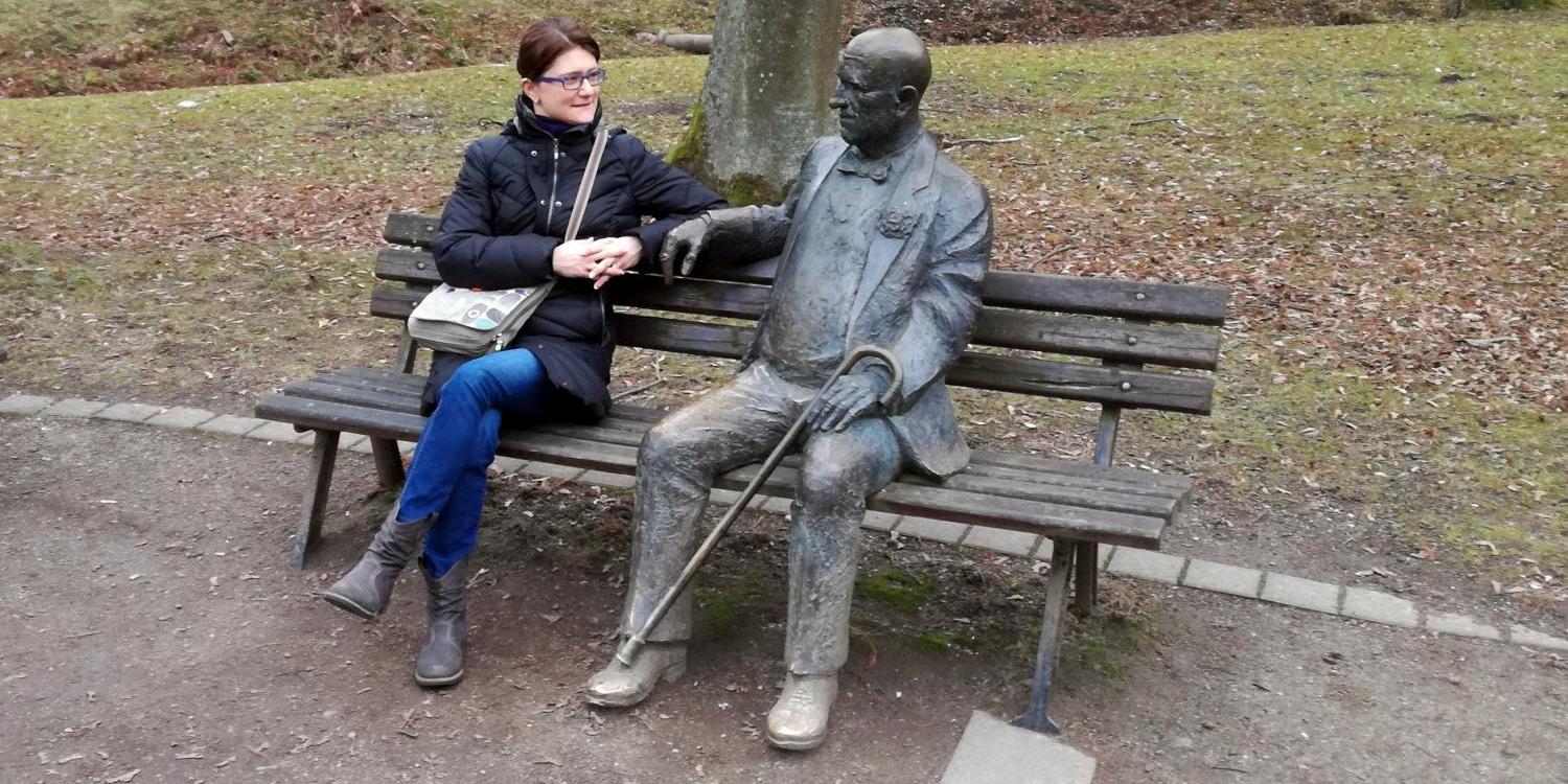 Frau sitzt neben einer Statue auf einer Parkbank, herbstliche Umgebung.