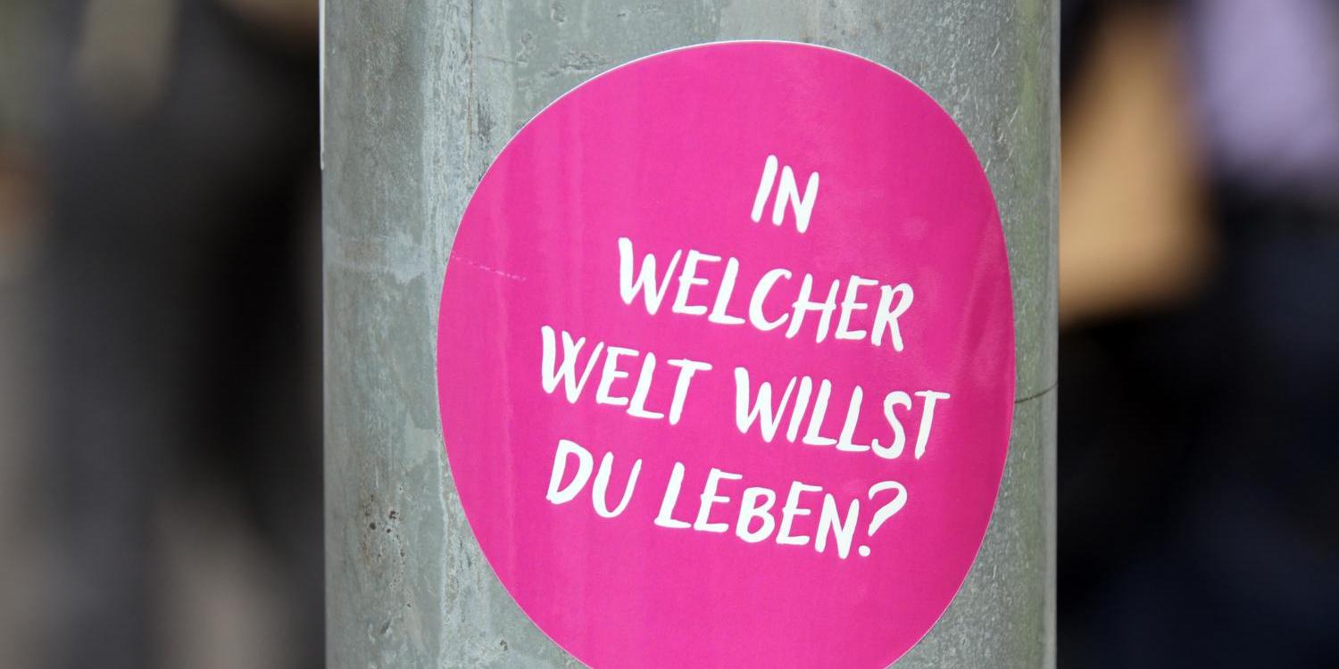 Das Foto zeigt einen runden, pinken Aufkleber auf einem grauen Metallpfosten mit der weißen Aufschrift: „In welcher Welt willst du leben?'