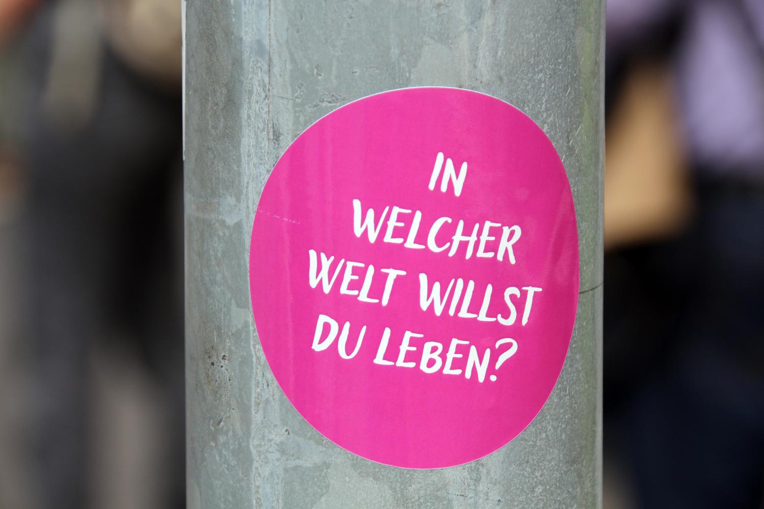 Das Foto zeigt einen runden, pinken Aufkleber auf einem grauen Metallpfosten mit der weißen Aufschrift: „In welcher Welt willst du leben?'