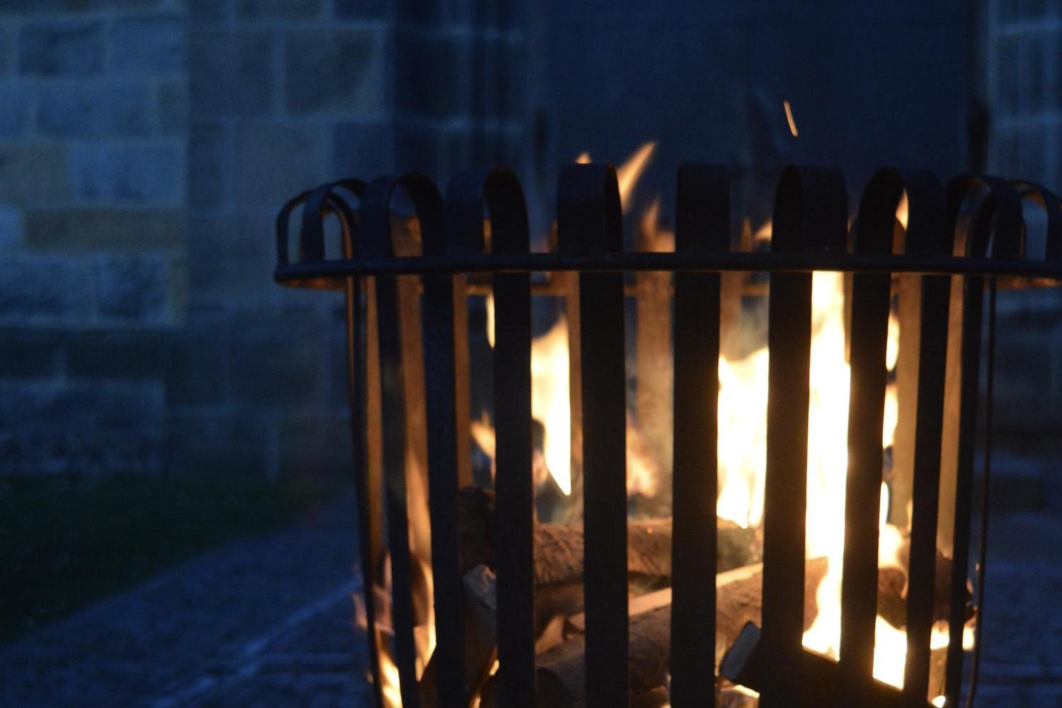 Ein offenes Feuer brennt in einem Metallkorb im Freien; die Flammen leuchten warm in der Dunkelheit vor einer steinernen Gebäudewand.