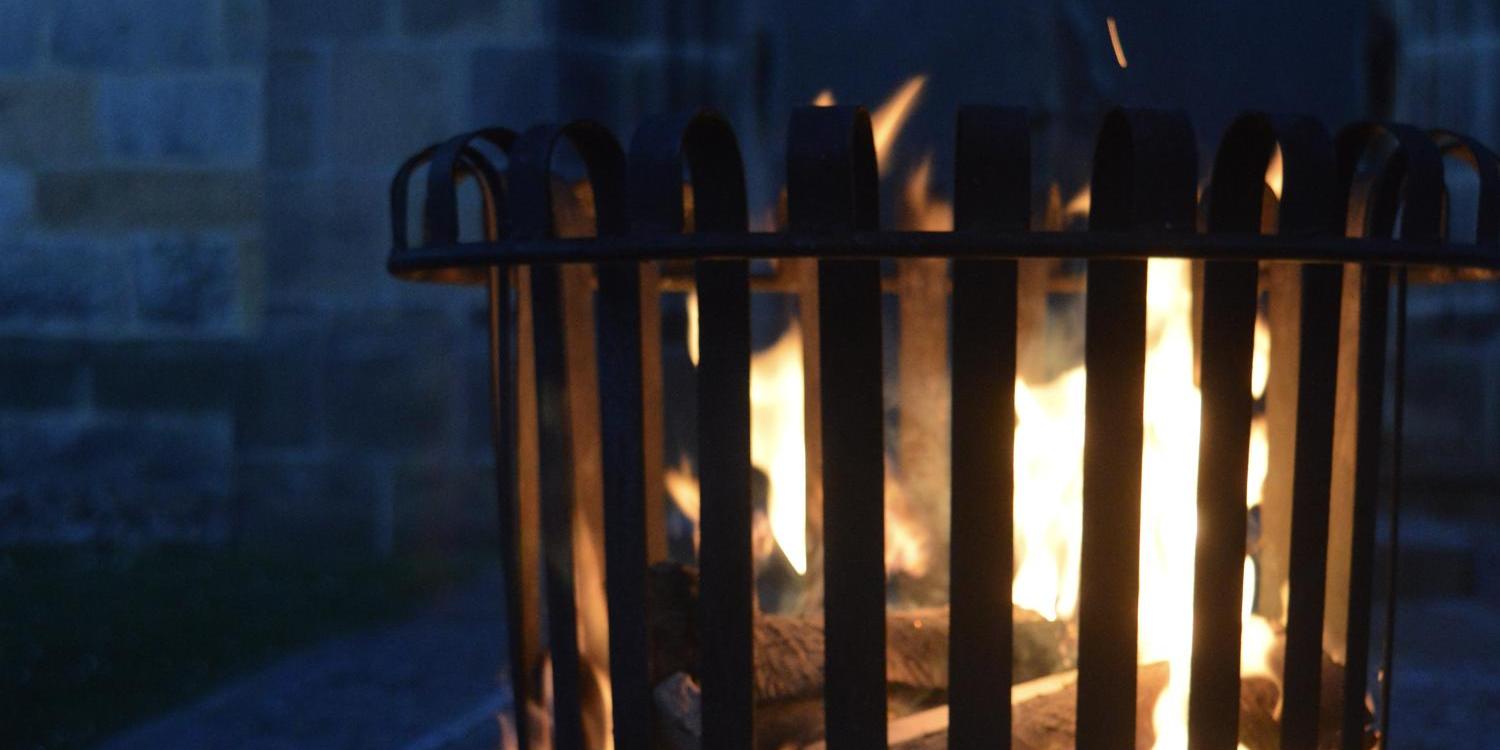 Ein offenes Feuer brennt in einem Metallkorb im Freien; die Flammen leuchten warm in der Dunkelheit vor einer steinernen Gebäudewand.
