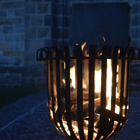 Ein offenes Feuer brennt in einem Metallkorb im Freien; die Flammen leuchten warm in der Dunkelheit vor einer steinernen Gebäudewand.