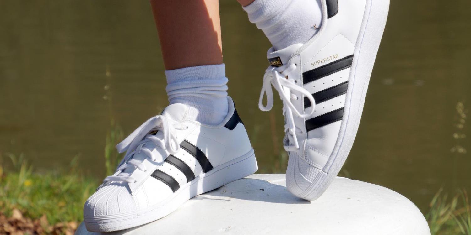 Weiße Turnschuhe an Füßen, die auf einem runden Stein am Wasser stehen.