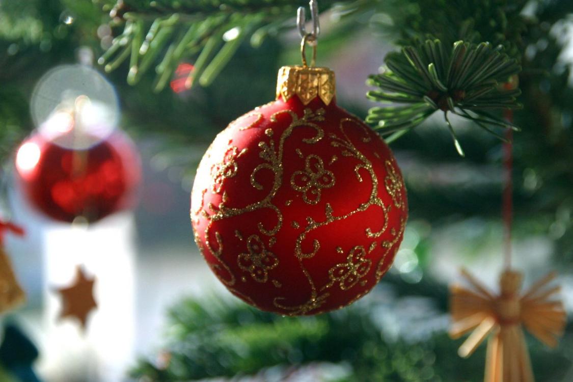 Das Foto zeigt eine rote Christbaumkugel mit goldenen Ornamentmustern, die an einem grünen Tannenzweig hängt, während im Hintergrund weitere Weihnachtsdekorationen verschwommen sichtbar sind.