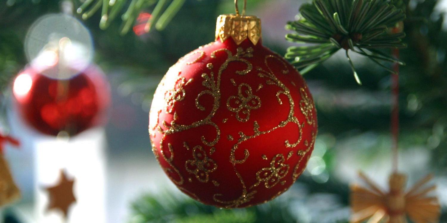 Das Foto zeigt eine rote Christbaumkugel mit goldenen Ornamentmustern, die an einem grünen Tannenzweig hängt, während im Hintergrund weitere Weihnachtsdekorationen verschwommen sichtbar sind.