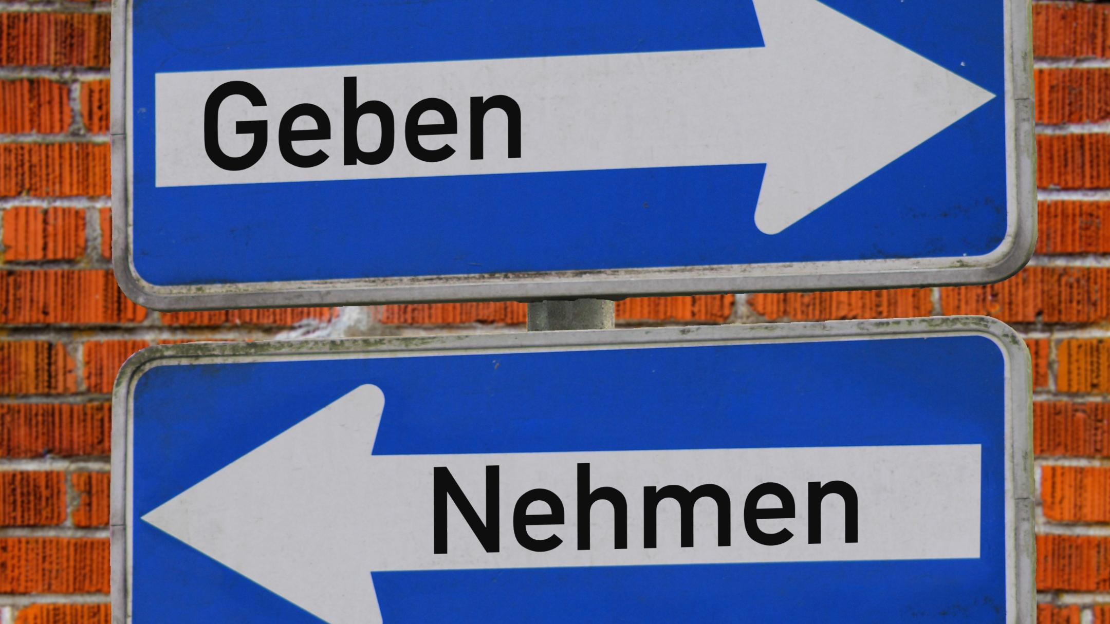 Das Foto zeigt zwei blaue Verkehrsschilder vor einer Backsteinmauer – eines mit einem weißen Pfeil nach rechts und dem Wort „Geben“, das andere mit einem weißen Pfeil nach links und dem Wort „Nehmen“.