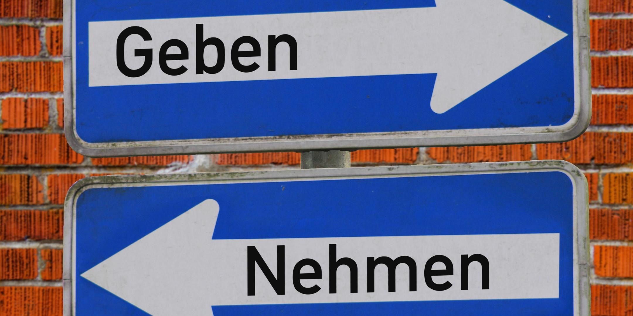 Das Foto zeigt zwei blaue Verkehrsschilder vor einer Backsteinmauer – eines mit einem weißen Pfeil nach rechts und dem Wort „Geben“, das andere mit einem weißen Pfeil nach links und dem Wort „Nehmen“.